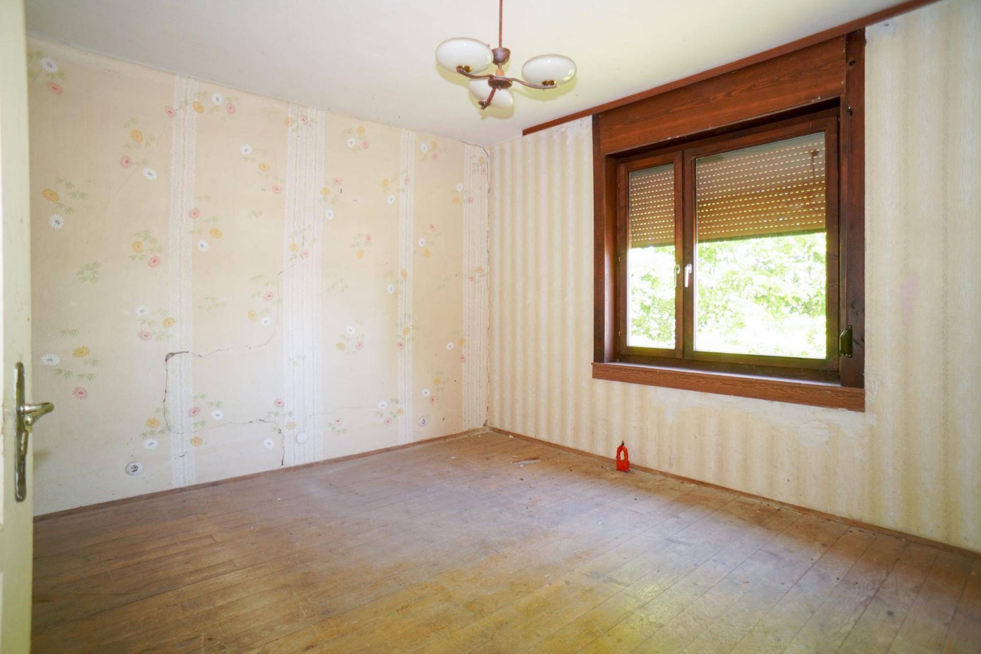 Schlafzimmer mit floraler Tapete, Holzfensterrahmen und Renovierungsbedarf.