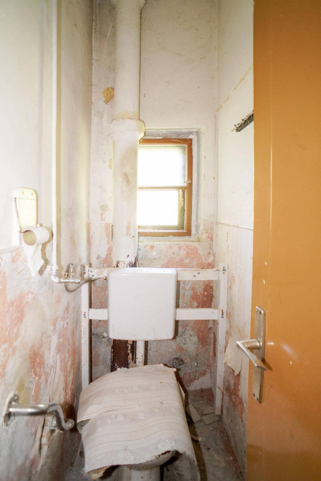 Separate Toilette mit Fenster und sichtbaren Installationsrohren, die renovierungsbedürftig ist.
