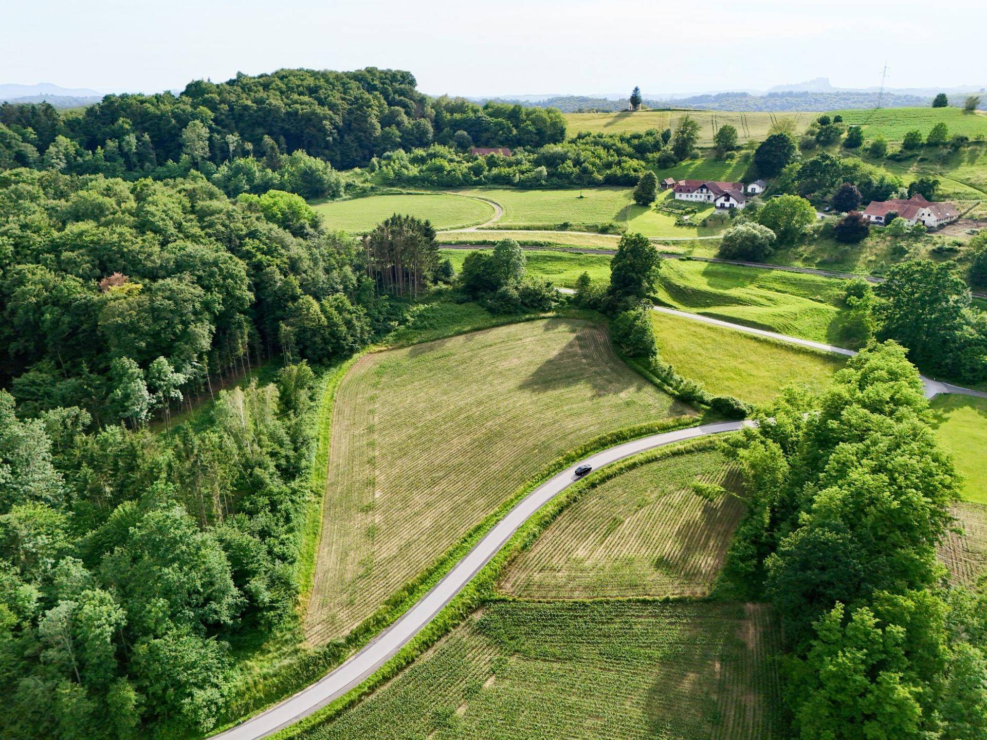 Weitläufige ländliche Szenerie mit Feldern, Wäldern und einer kurvigen Straße, auf der ein Auto fährt.