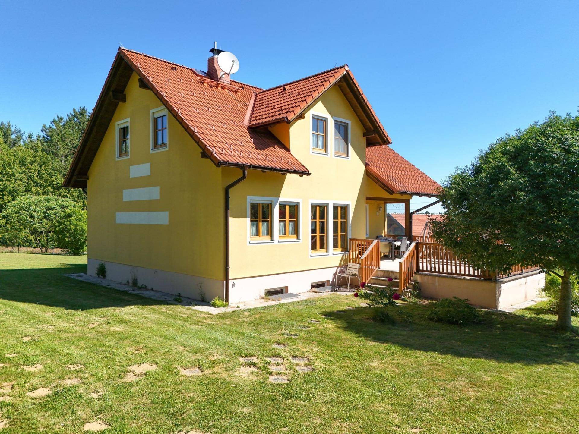 Seitenansicht des gelben Hauses mit rotem Ziegeldach, umgeben von einem gepflegten Rasen und hohen Bäumen.
