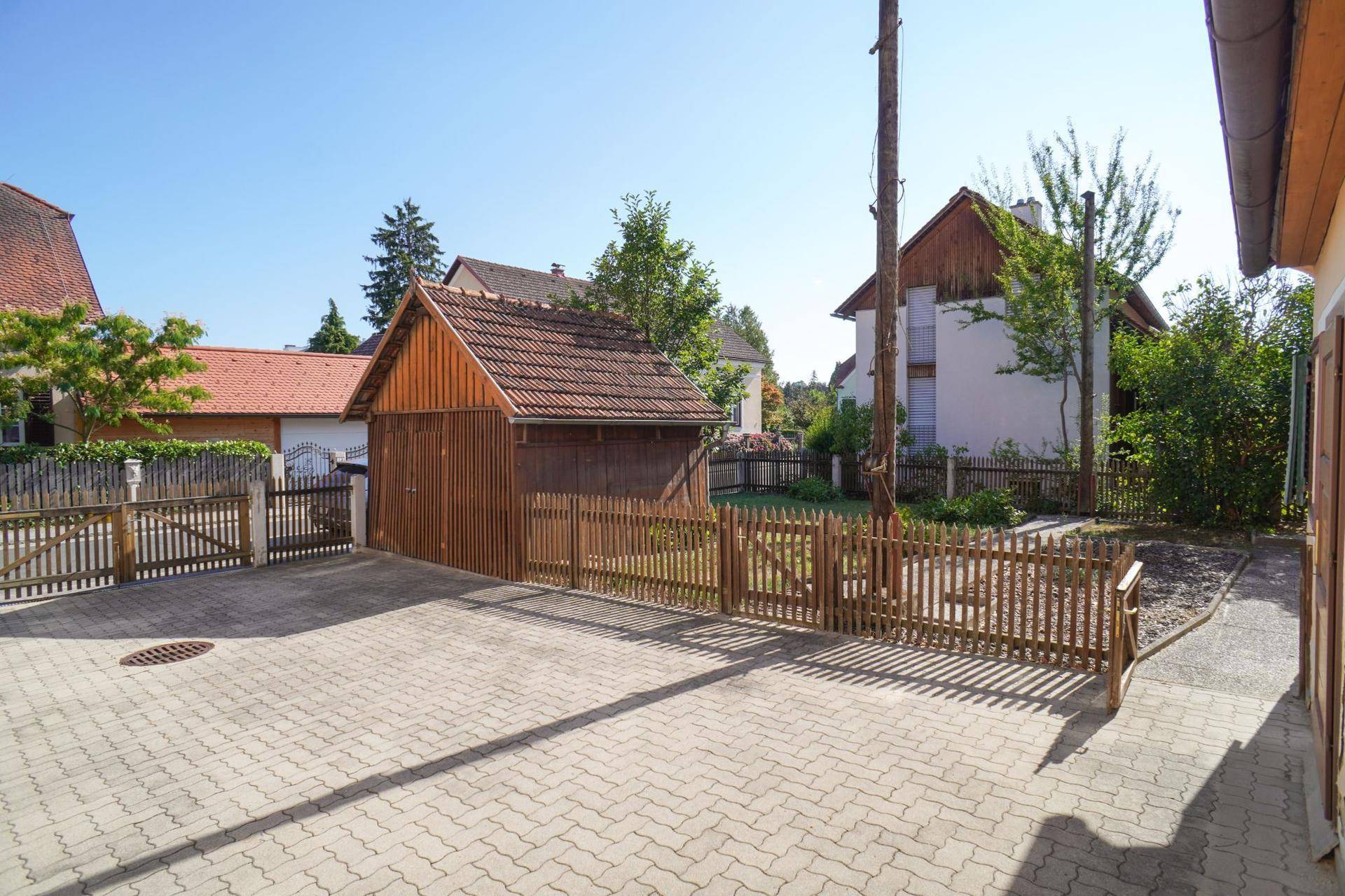 Gepflasterte Einfahrt mit Holzzaun und einer Holzgarage, umgeben von Grünflächen.