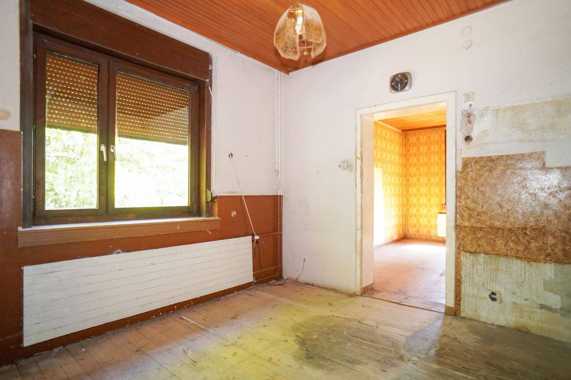Raum mit Holzdecke, teilweise entfernter Tapete und Blick in einen weiteren Raum.