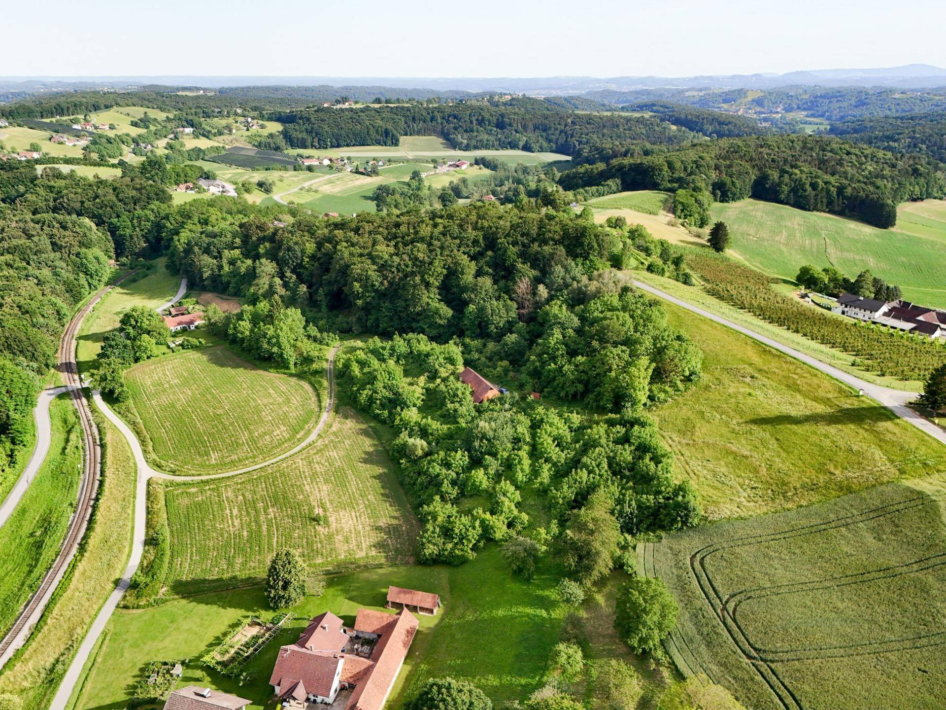 Panoramablick auf eine grüne Hügellandschaft mit Feldern, Wäldern und einer Bahnlinie in der Ferne.