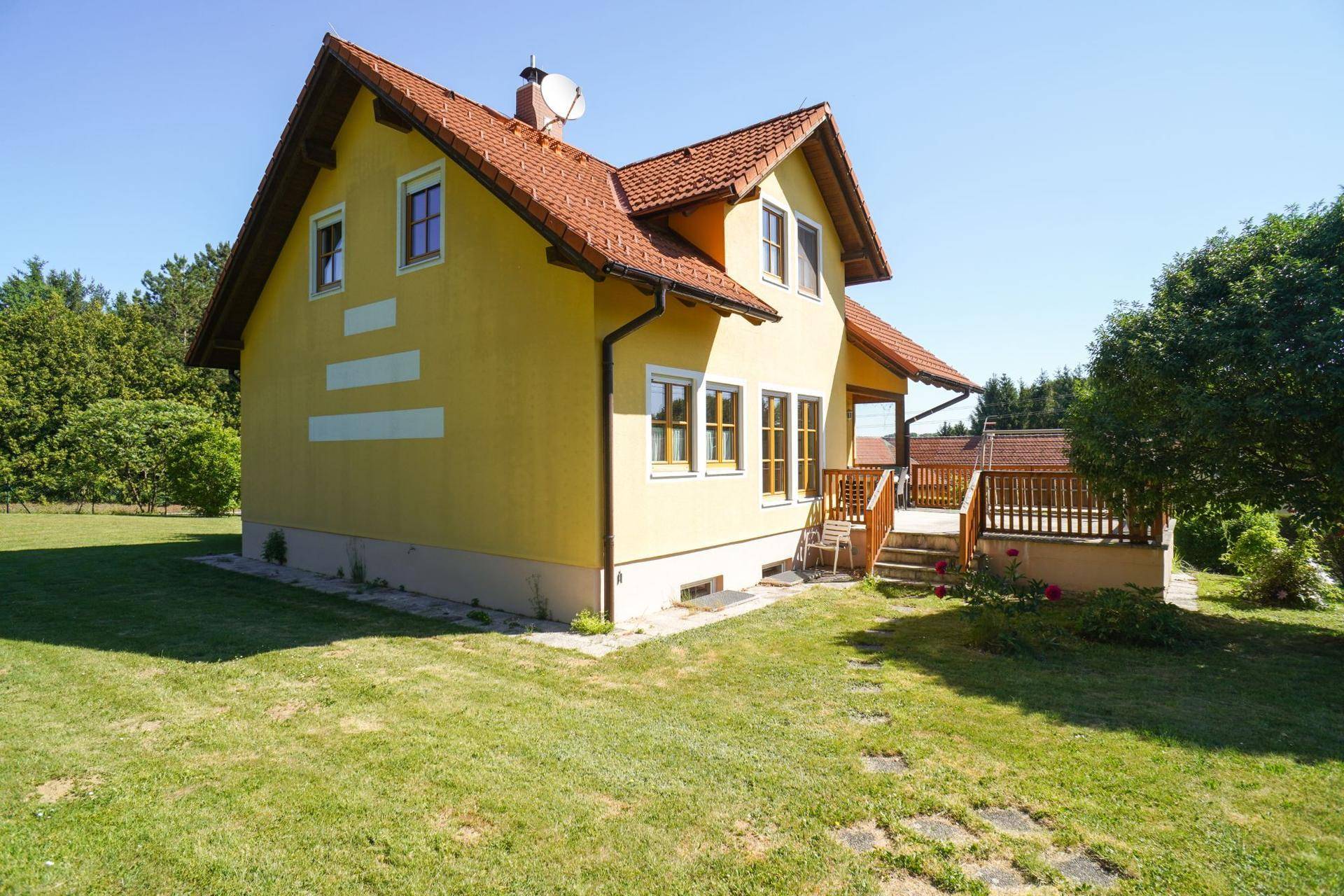 Gelbes Einfamilienhaus mit rotem Ziegeldach und einer Holzterrasse, umgeben von grünem Rasen.