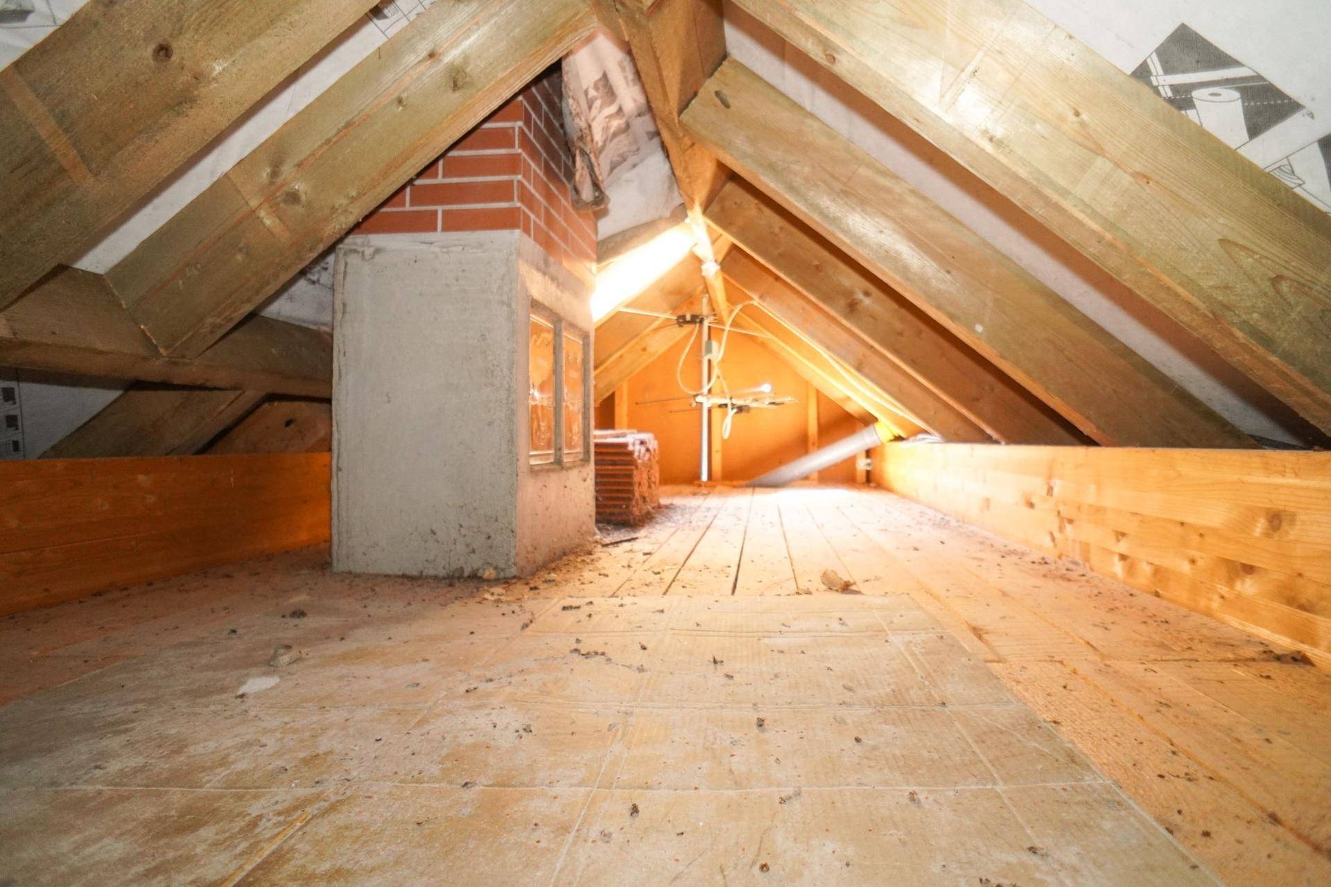 Unausgebauter Dachboden mit sichtbaren Holzbalken und einem gemauerten Kamin, bietet Ausbaupotenzial.