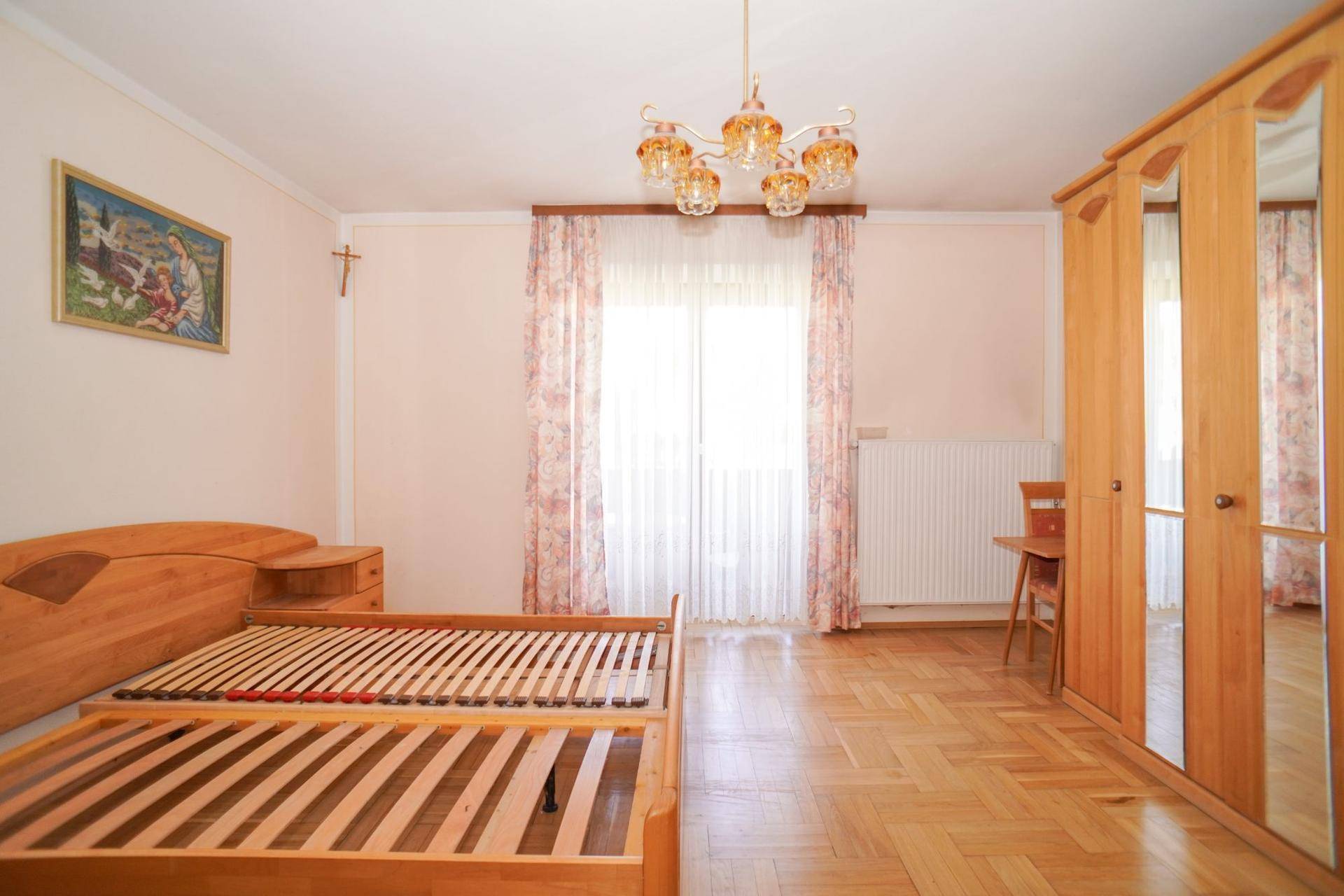 Helles Schlafzimmer mit Parkettboden, Holzmöbeln und einem großen Fenster mit Vorhängen.