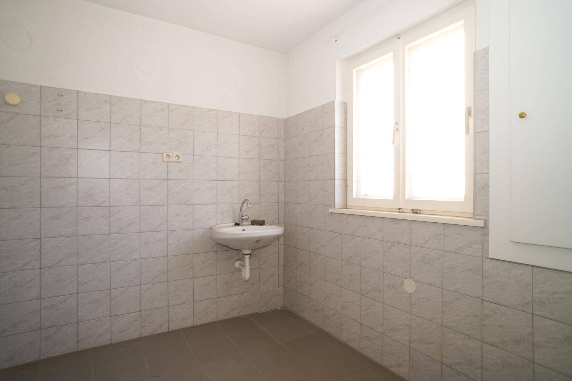 Helles Badezimmer mit hellgrauen Fliesen, kleinem Waschbecken und Fenster.