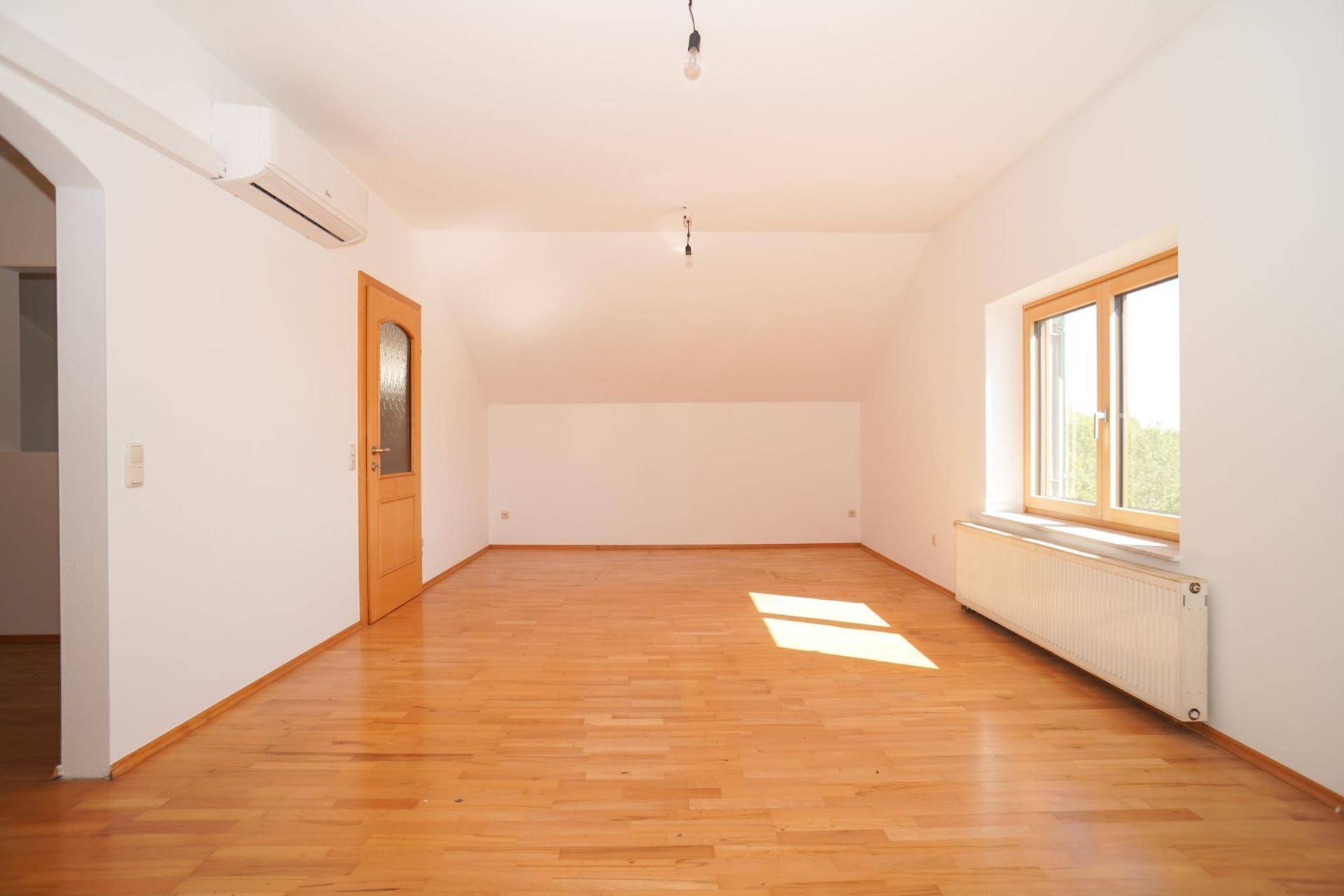 Geräumiges Dachzimmer mit Holzboden, Fenster und Klimaanlage unter der Dachschräge.