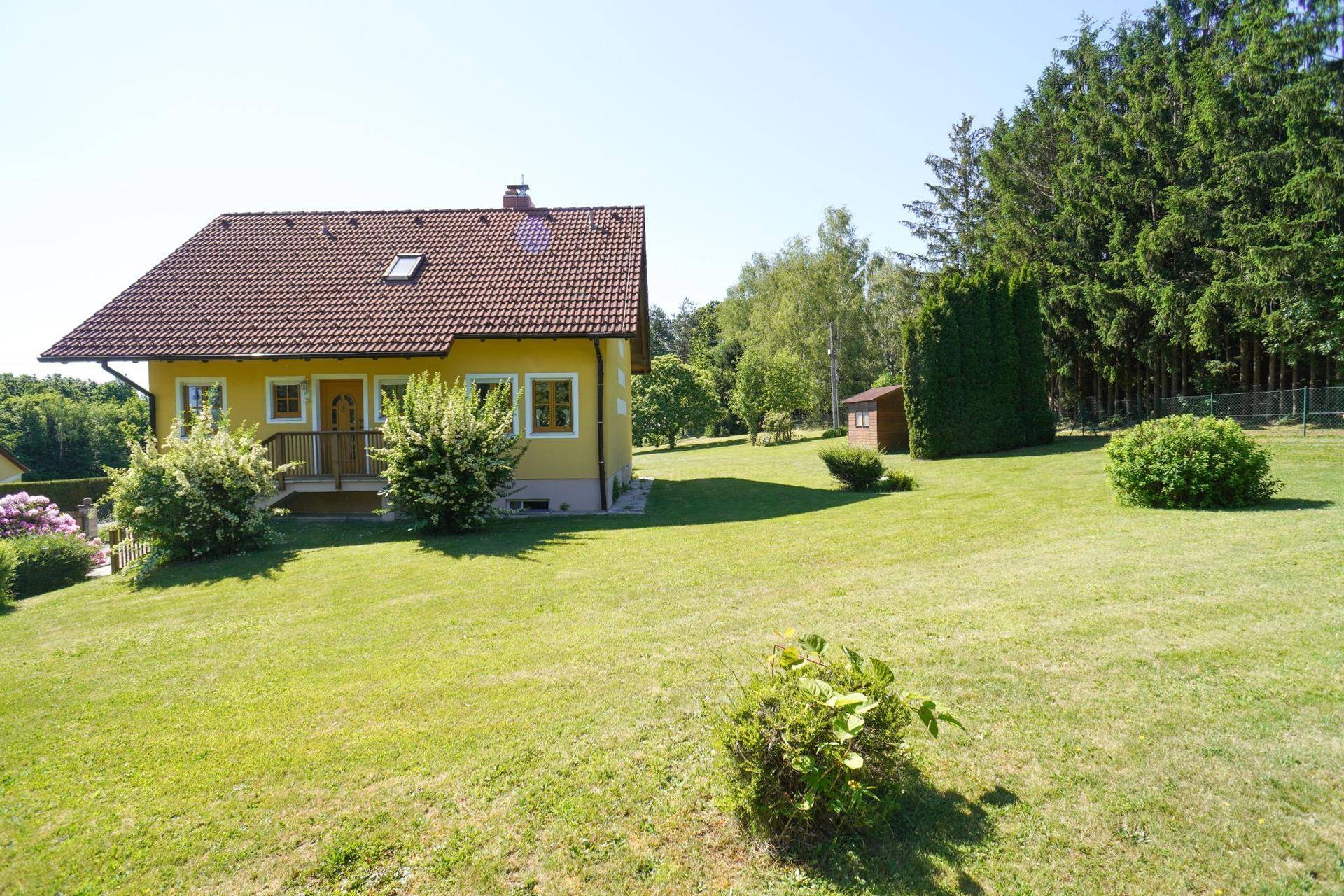 Vorderansicht des gelben Hauses mit weitläufigem Rasen und blühenden Sträuchern im Sonnenschein.