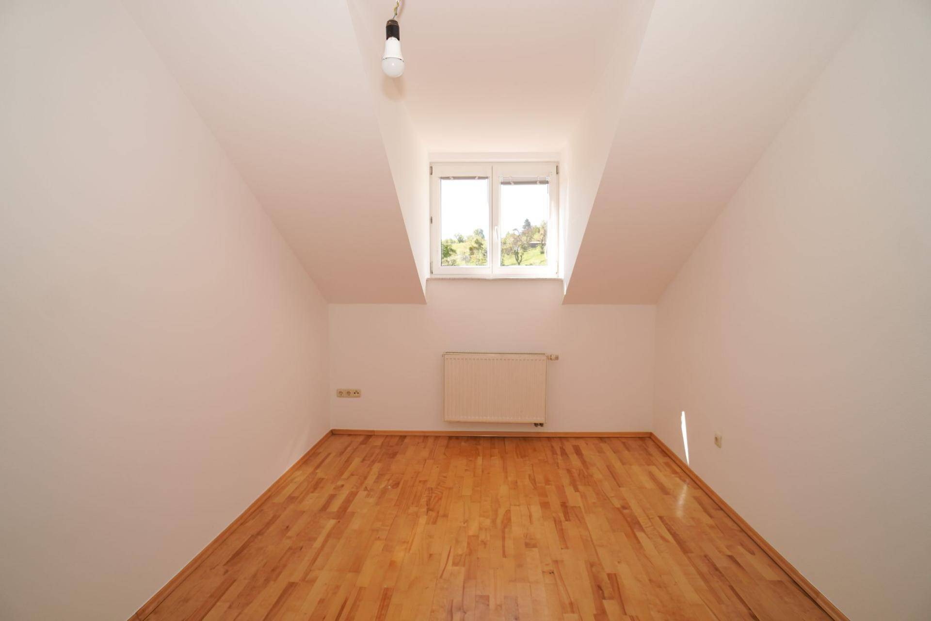 Helles Dachzimmer mit Holzboden und Fenster, das viel Tageslicht hereinlässt.