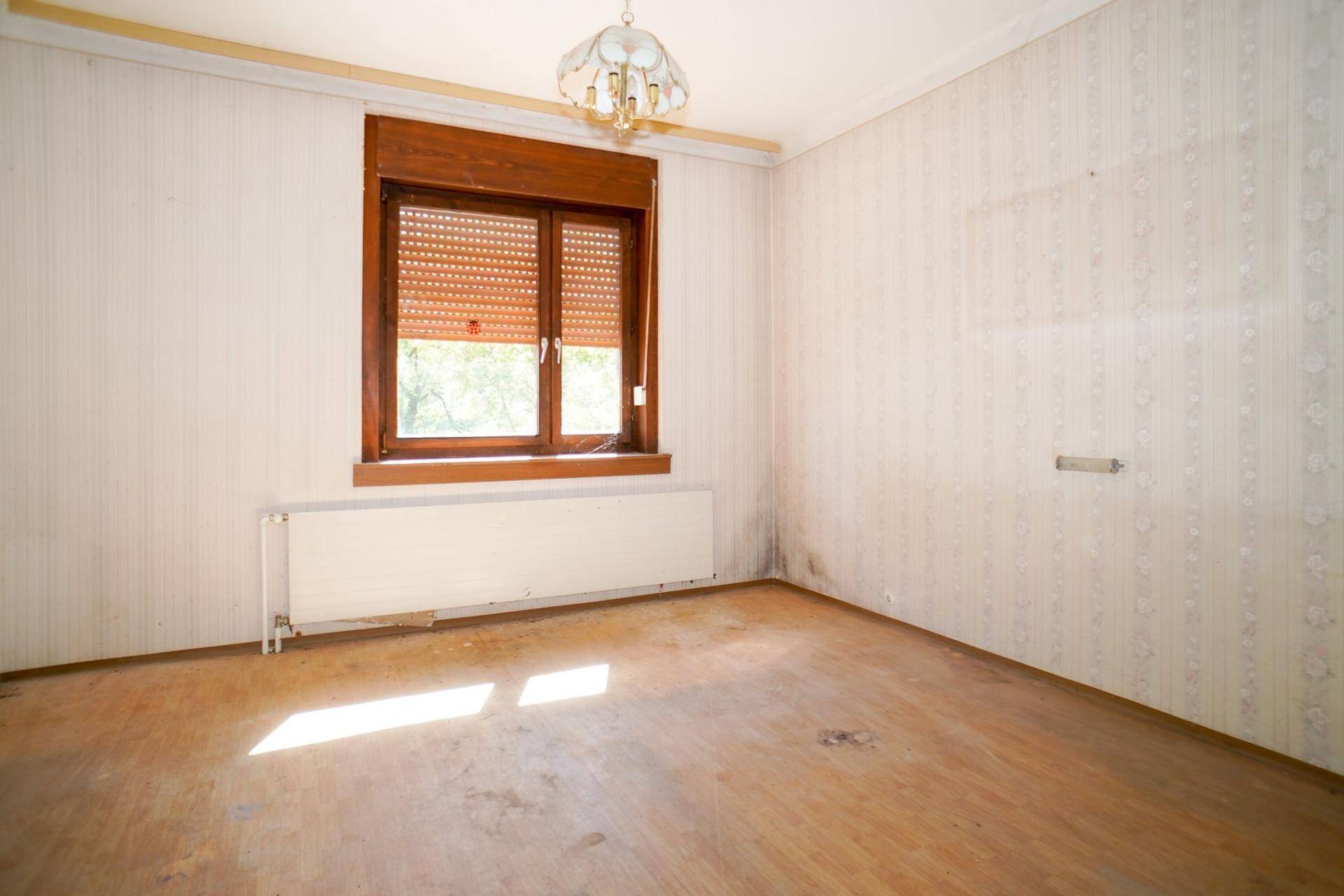 Schlafzimmer mit gestreifter Tapete, Holzfensterrahmen und Heizkörper, das Renovierungsbedarf zeigt.