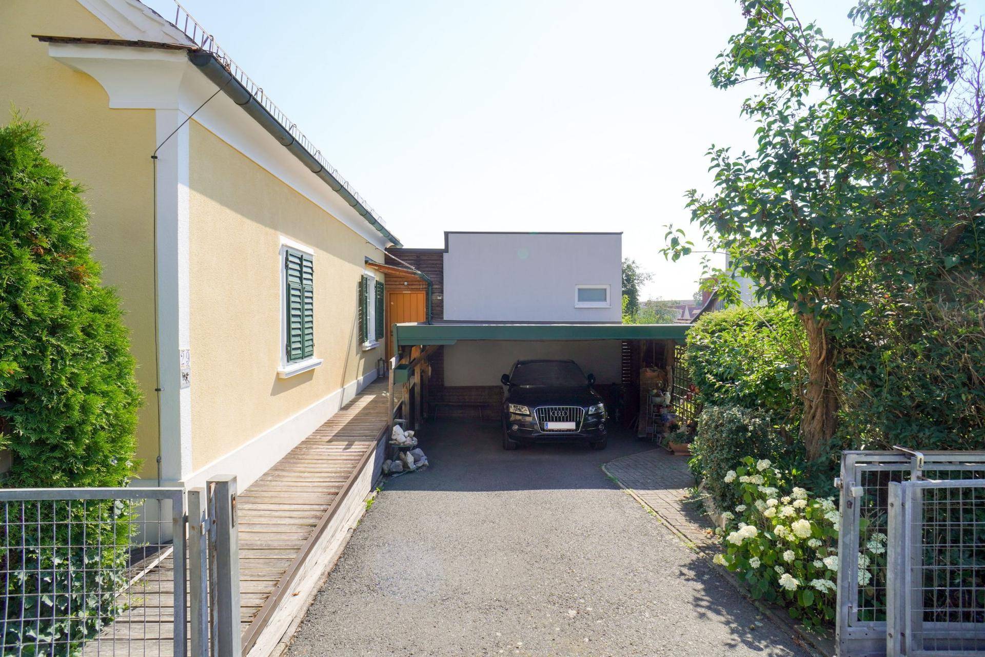 Einfahrt mit Carport und einem gelben Haus mit grünen Fensterläden, umgeben von viel Grün.