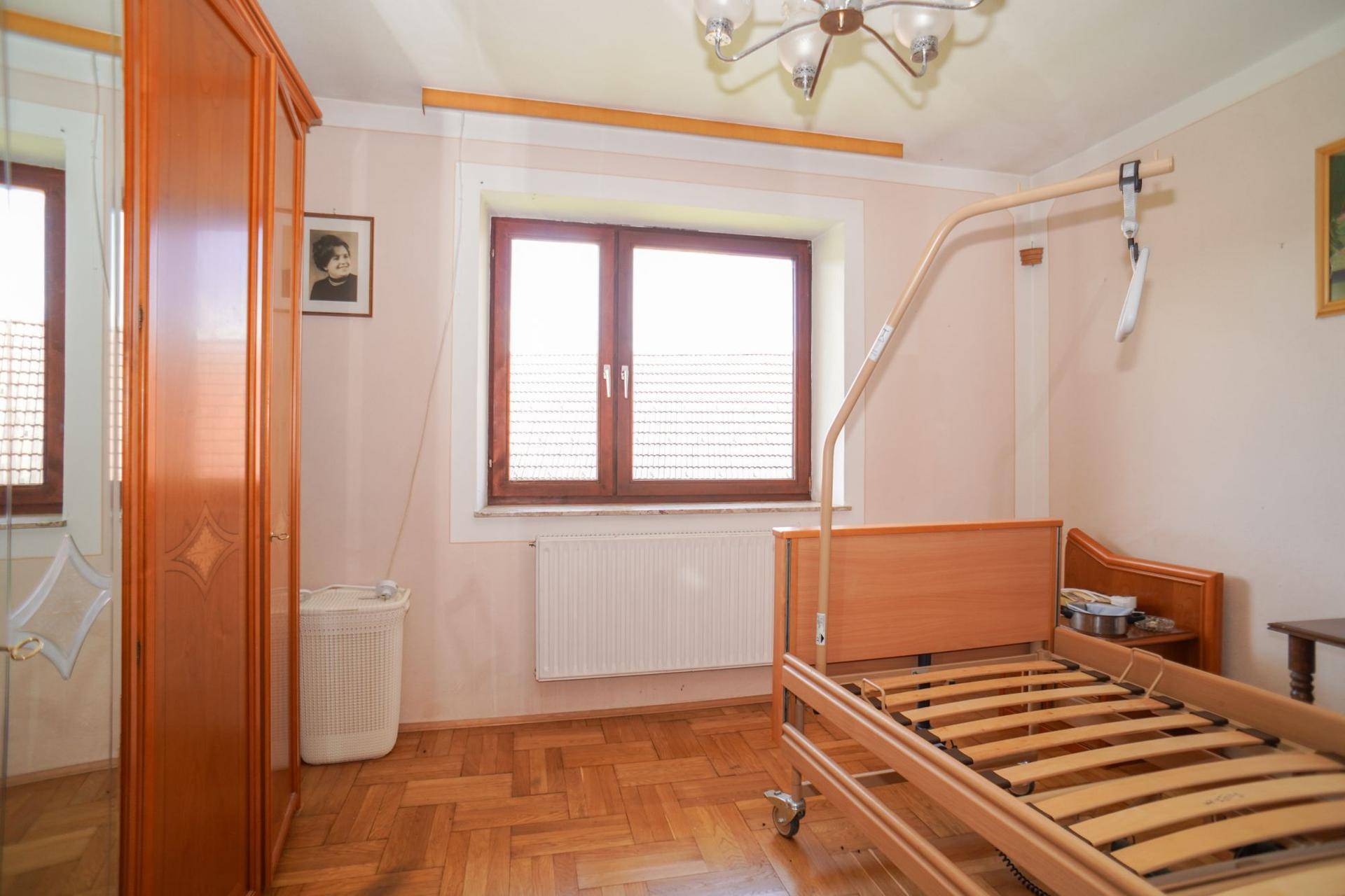 Schlafzimmer mit Parkettboden, einem Pflegebett und einem Fenster mit Jalousien.