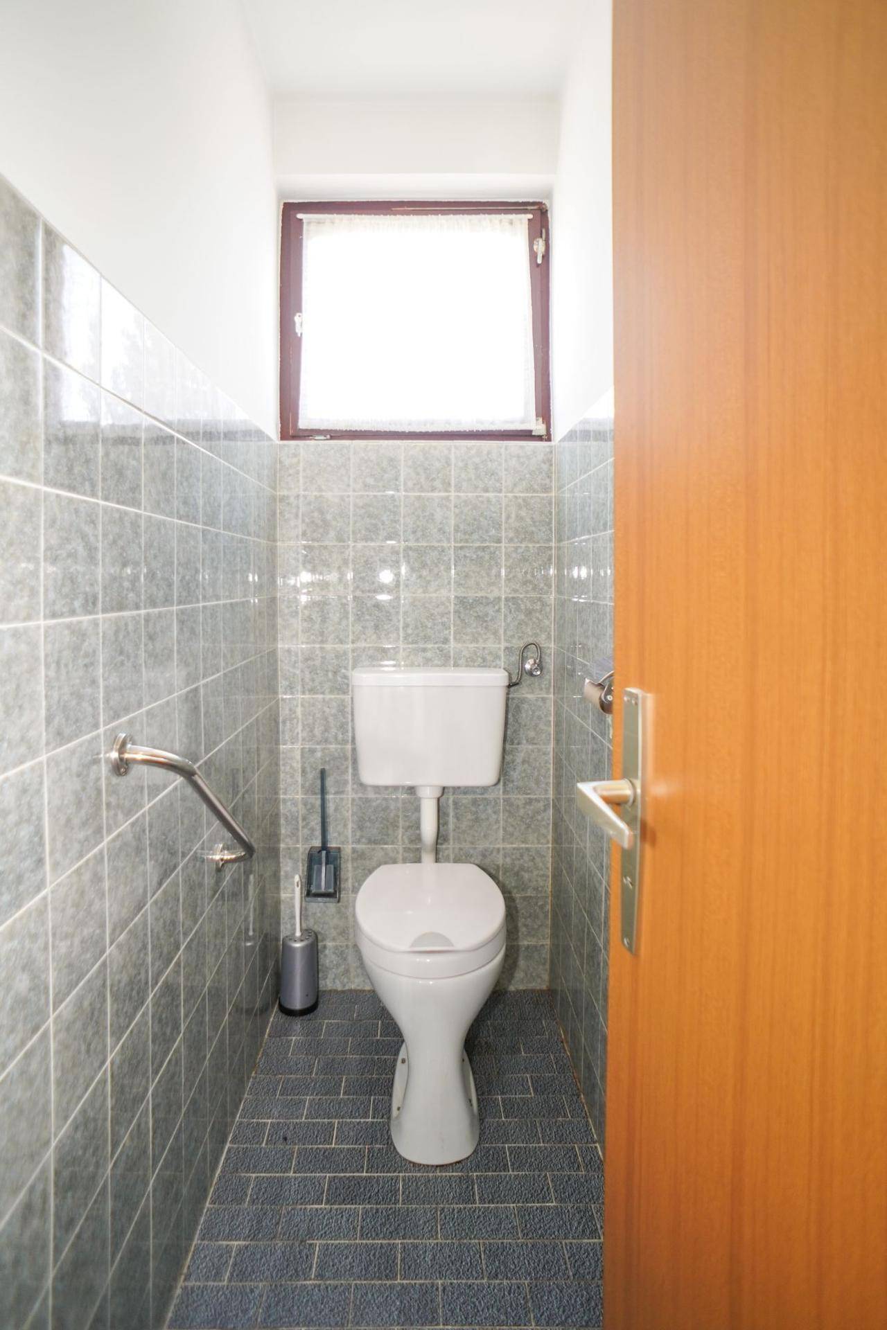 Separate Toilette mit grauen Fliesen, Fenster und Haltegriffen für Barrierefreiheit.