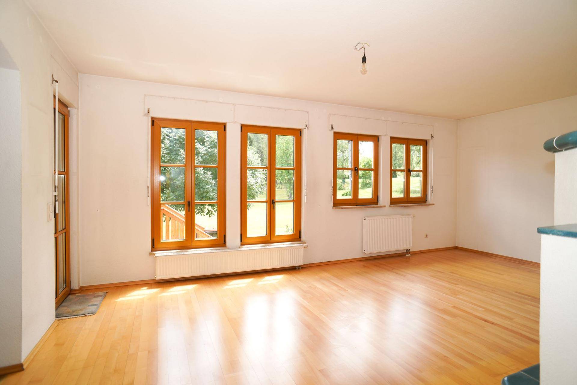 Helles Wohnzimmer mit Holzboden und Zugang zur Terrasse, umgeben von großen Fenstern mit grüner Aussicht.