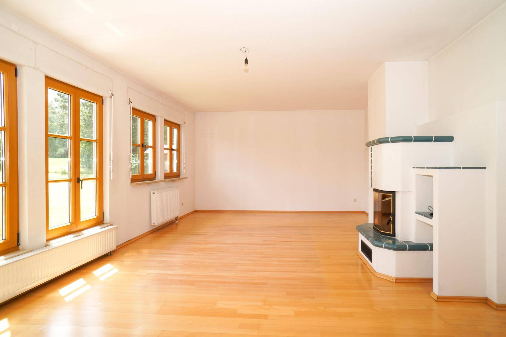 Geräumiges Wohnzimmer mit hellem Holzboden, großen Fenstern und einem traditionellen Kachelofen.