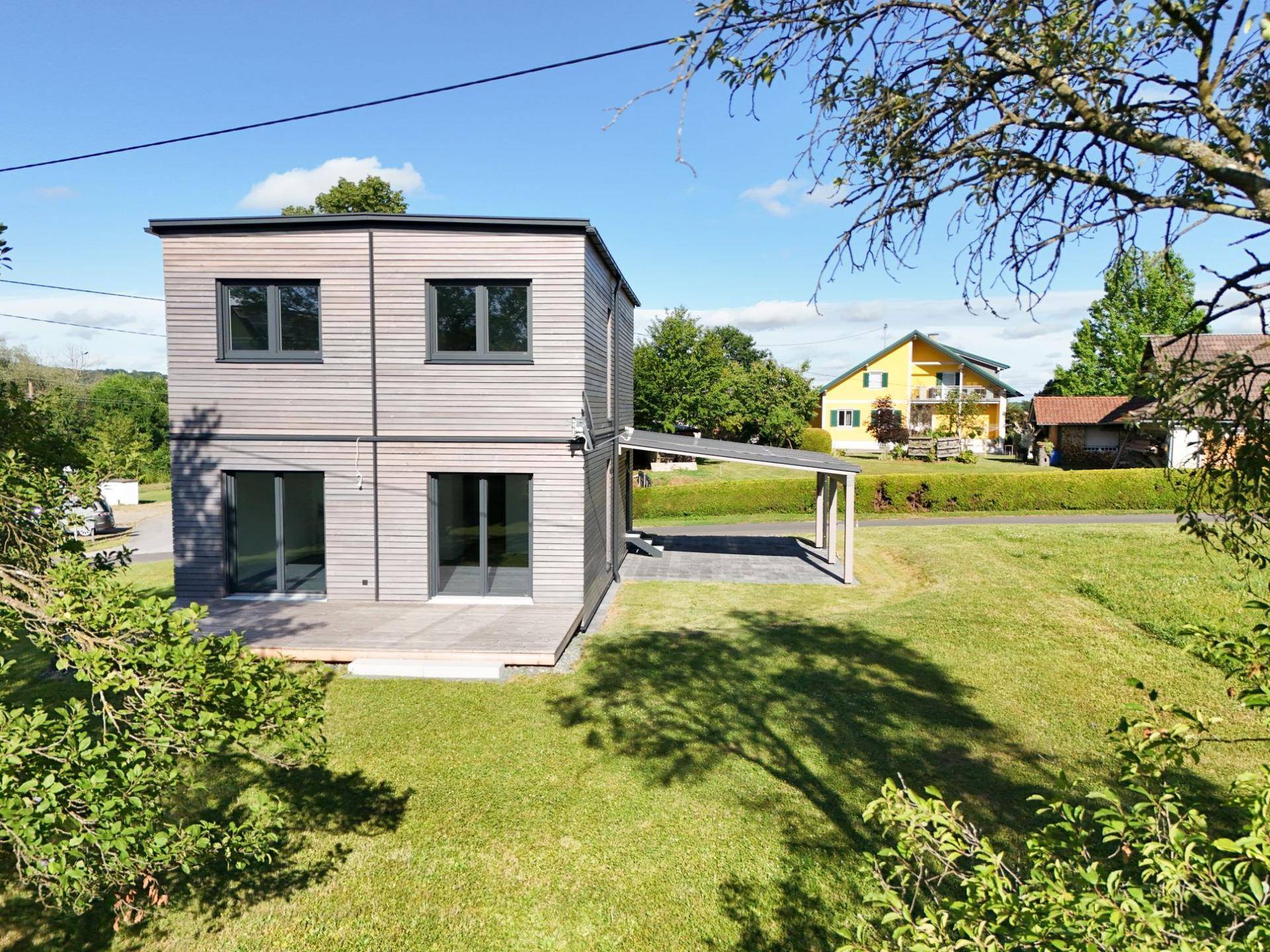 Modernes Einfamilienhaus mit Holzfassade, Carport und großem Garten in ländlicher Umgebung.