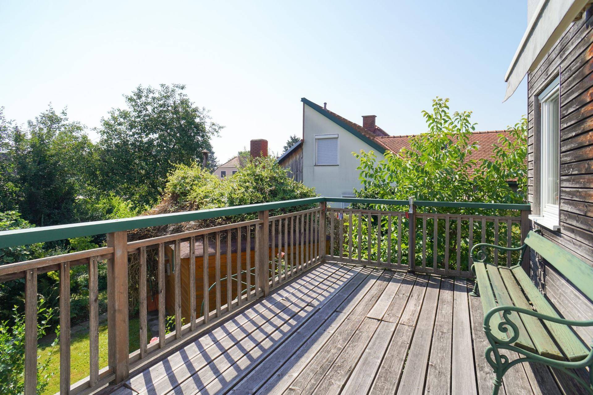 Geräumige Holzterrasse mit grüner Brüstung und Gartenbank, umgeben von Bäumen und viel Grün.