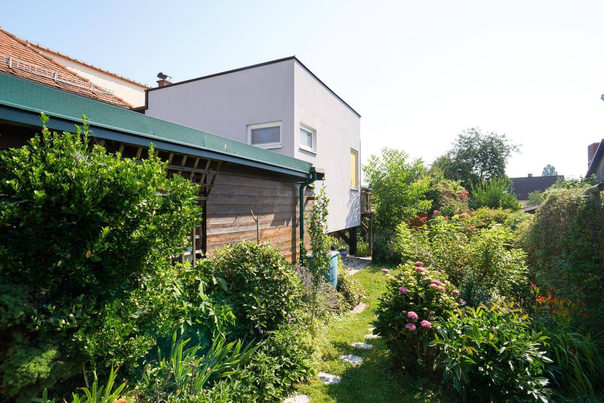 Verwunschener Gartenweg zwischen blühenden Büschen, der zu einem modernen Anbau führt.