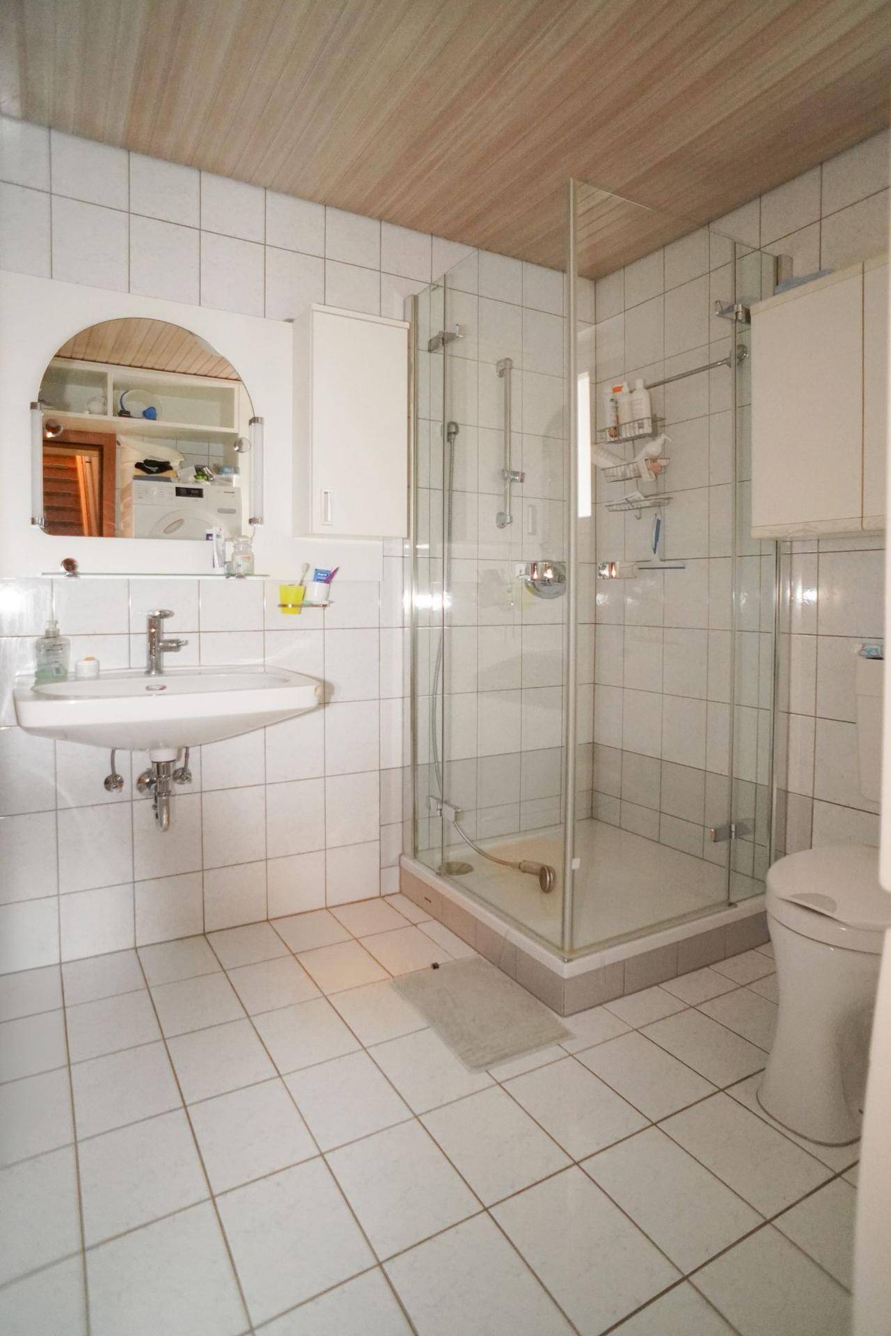 Badezimmer mit weißen Fliesen, Duschkabine aus Glas und einer Holzdecke.