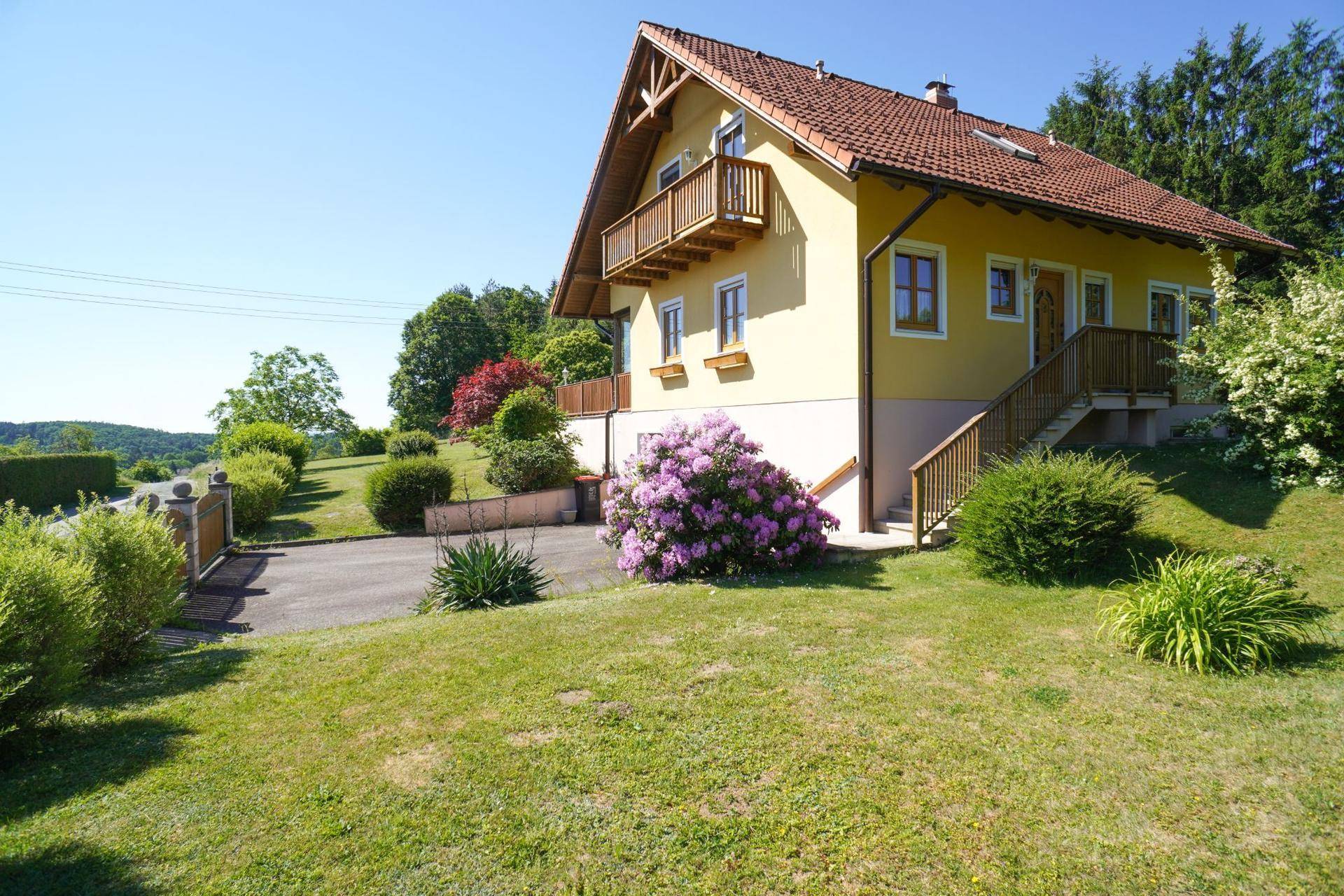 Vorderseite des Hauses mit Zufahrt, kleinem Holzbalkon und blühenden Rhododendren.
