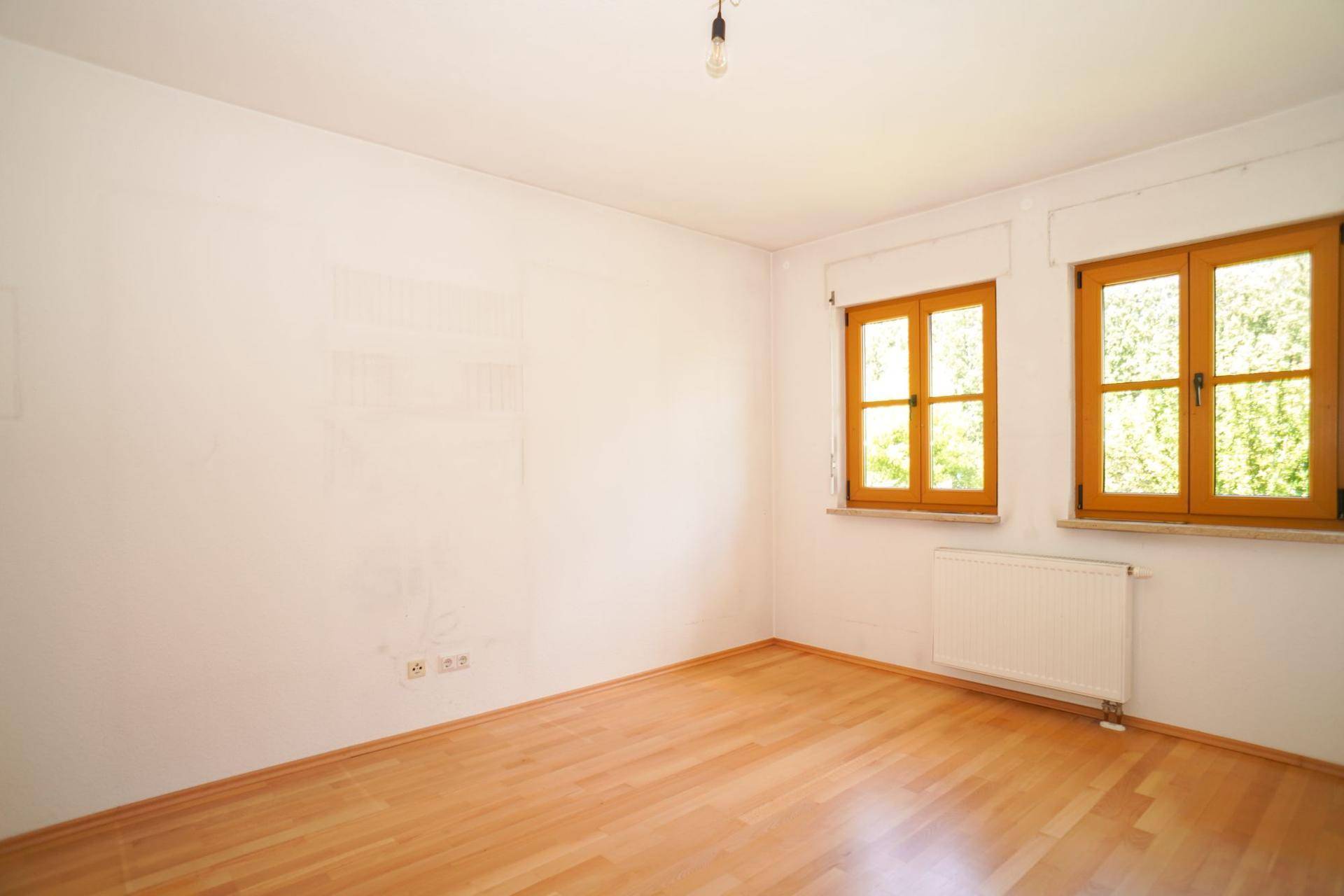 Helles Schlafzimmer mit Holzboden, weißen Wänden und zwei Fenstern mit Blick ins Grüne.