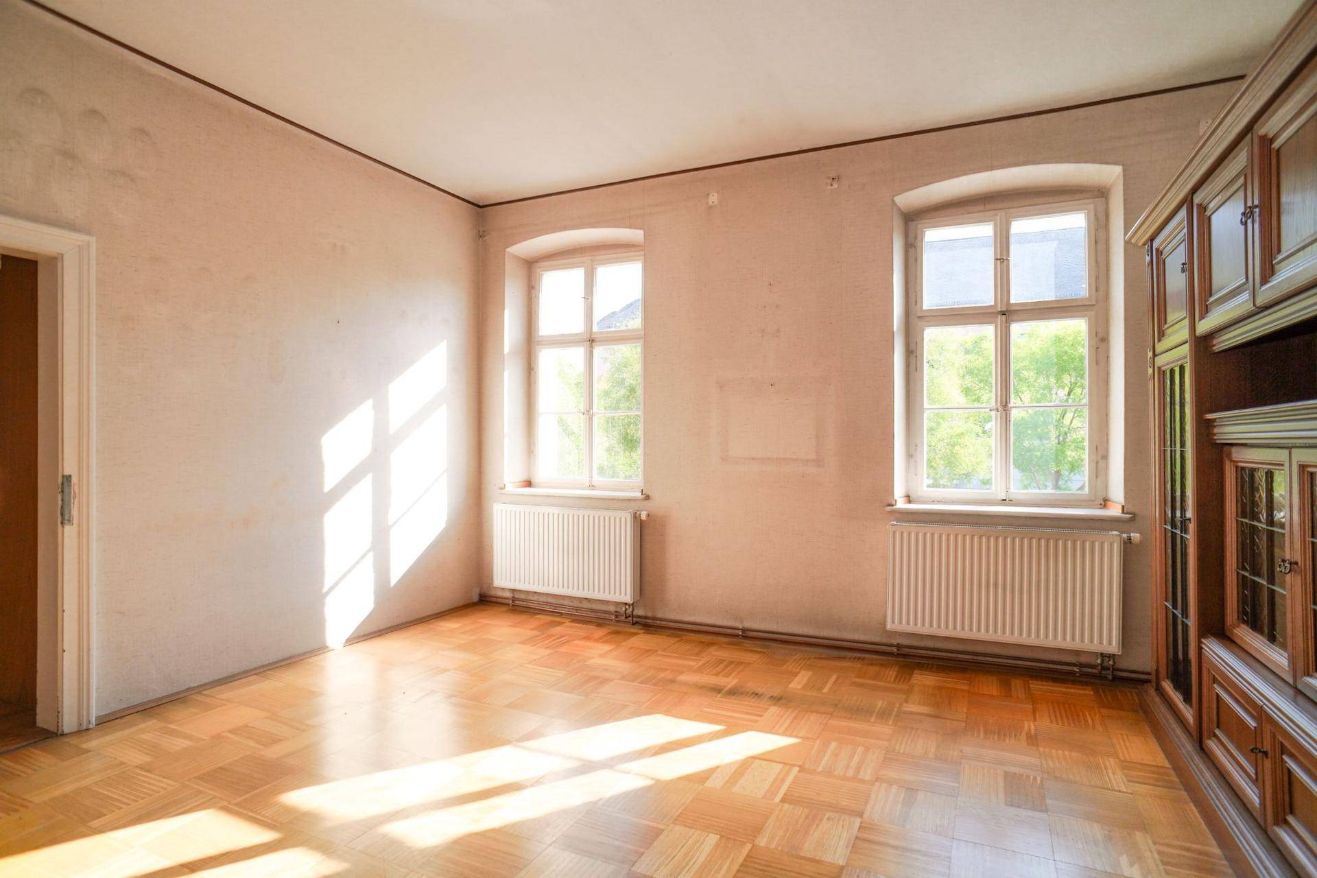 Helles Wohnzimmer mit klassischem Parkettboden und zwei großen Fenstern, sonnendurchflutet.