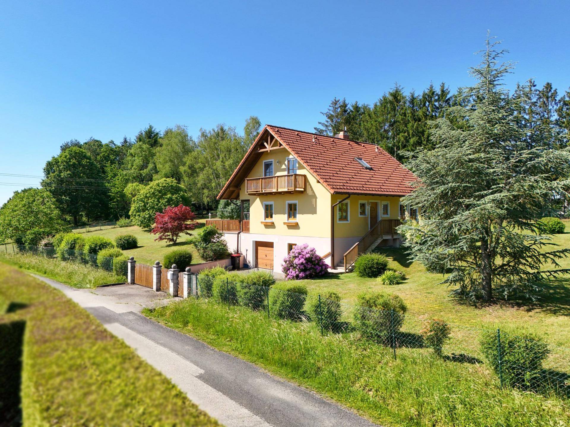Freistehendes Haus mit gelber Fassade und rotem Dach, eingebettet in eine grüne Landschaft mit Bäumen und Hecken.