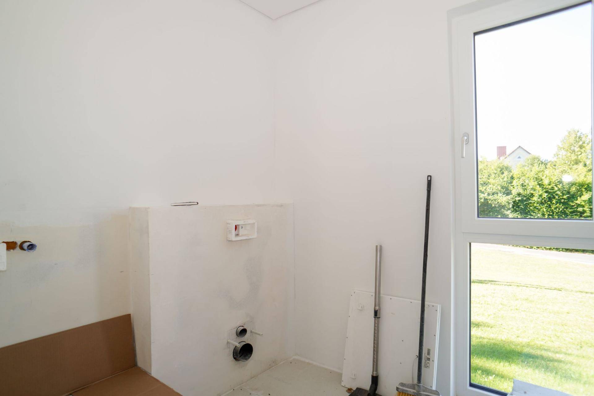 Unfertiger Raum mit Sanitäranschlüssen für ein WC und Fenster mit Blick ins Grüne.