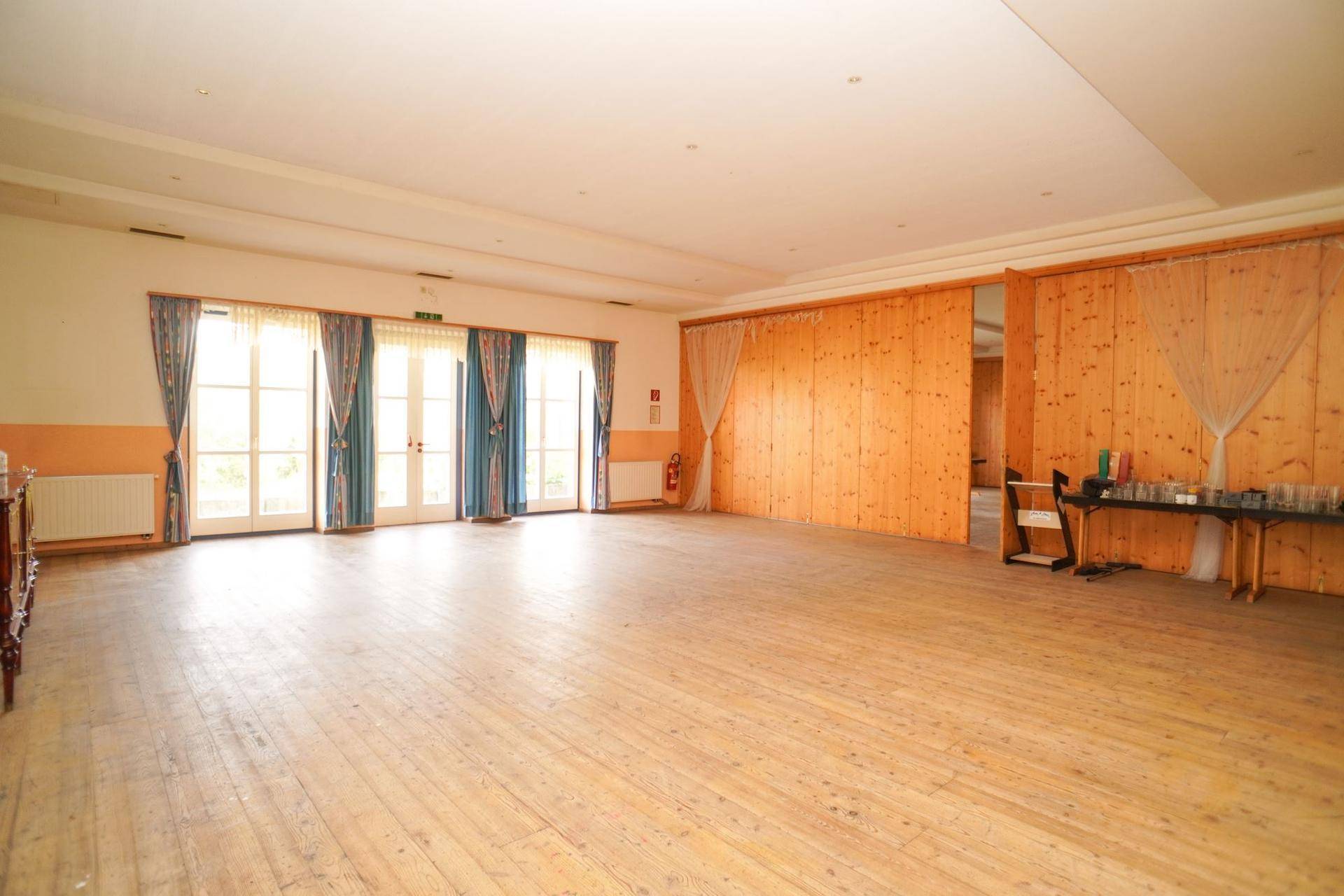 Großer Saal mit Holzdielenboden, Holzvertäfelung und Zugang zu mehreren Fenstertüren.