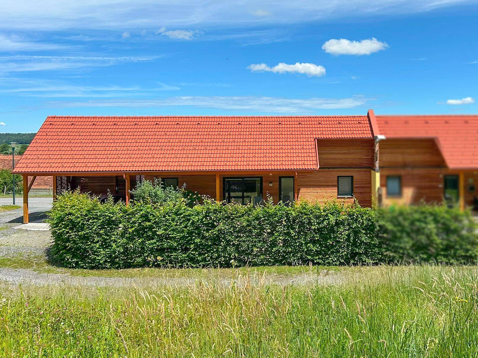 Langes Holzhaus mit rotem Ziegeldach und grüner Hecke vor einer weiten Wiese.