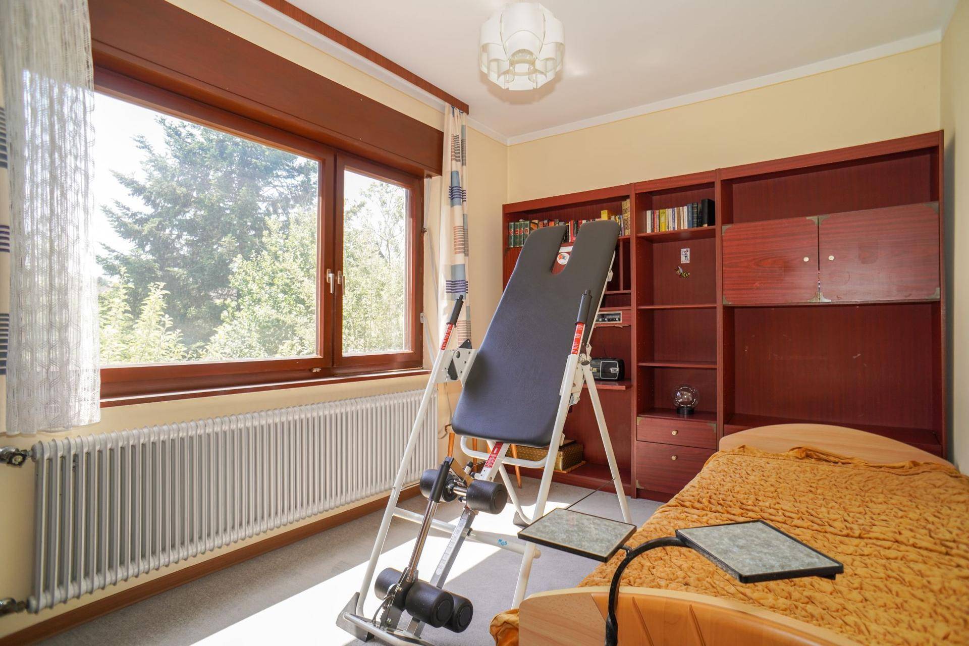 Vielseitiges Zimmer mit Bett, Bücherregal und großem Fenster mit Blick ins Grüne.