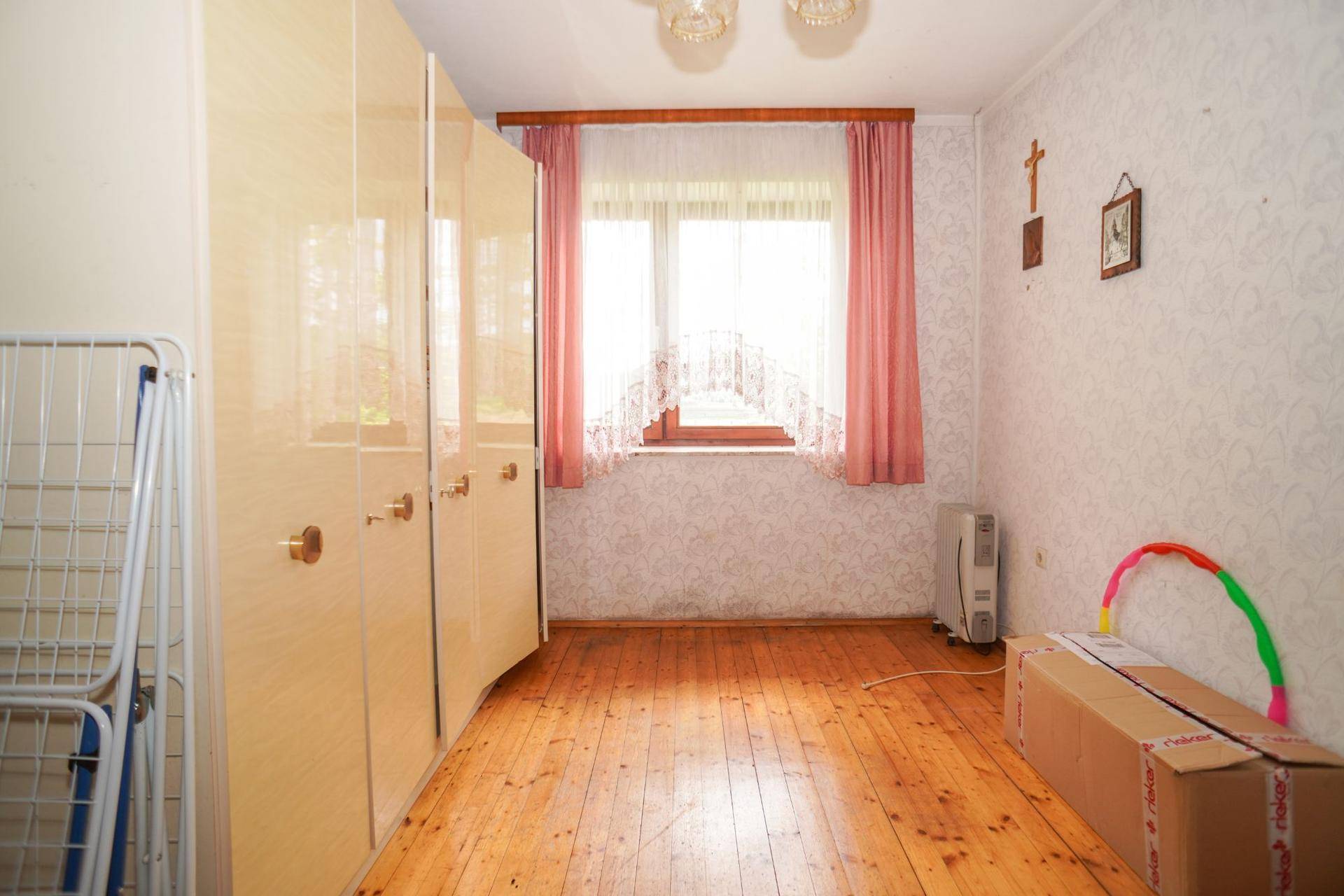 Schlafzimmer mit Holzdielenboden, gemusterter Tapete und einem Fenster mit rosa Vorhängen.