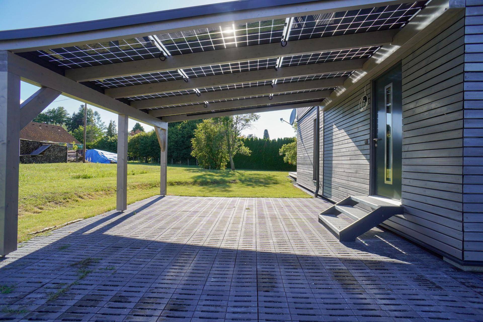 Überdachter Eingangsbereich mit Photovoltaikanlage auf dem Dach und gepflastertem Boden im Sonnenlicht.