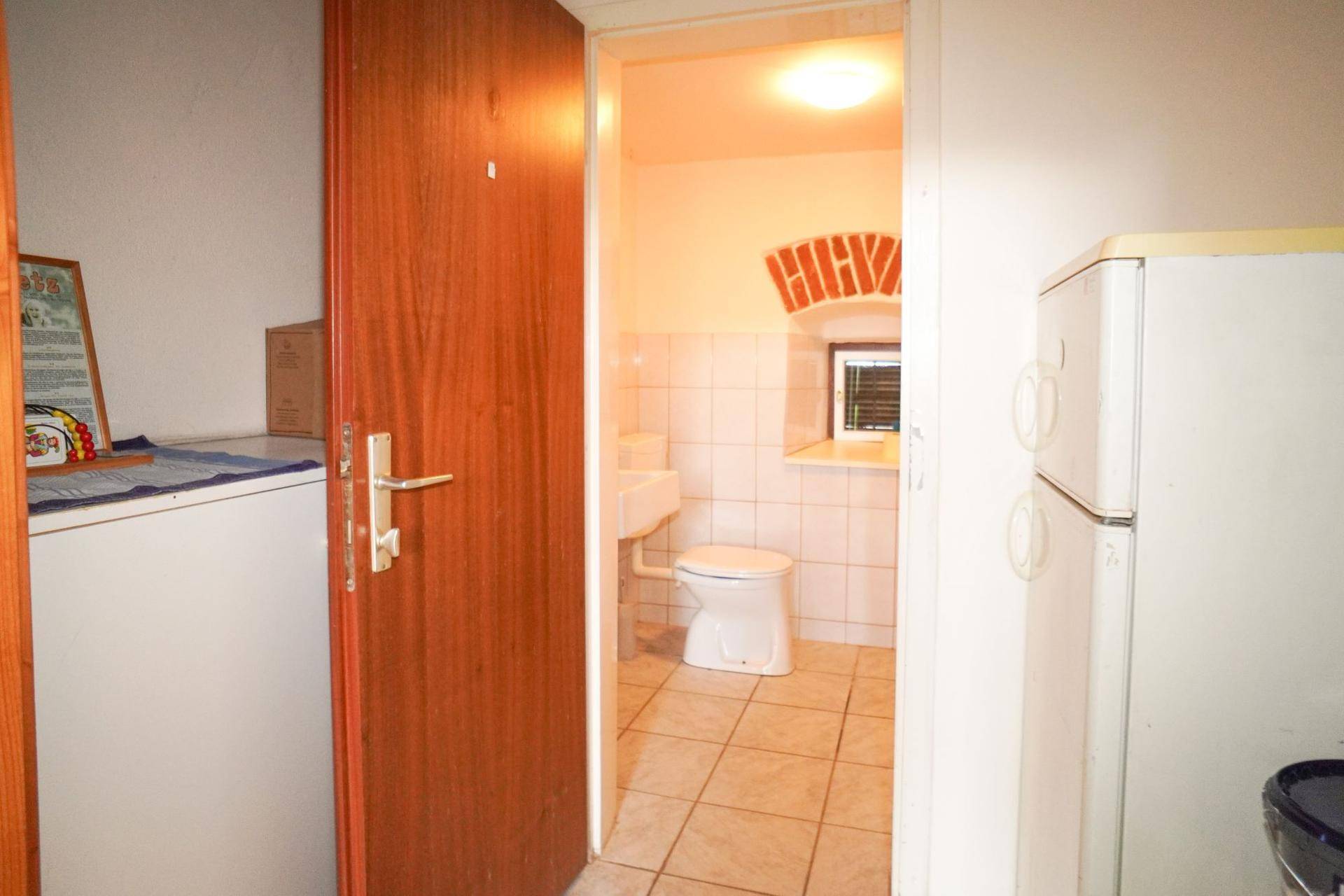 Kleines WC mit Fliesenboden und Fenster, zugänglich durch eine Holztür und Kühlschrank.