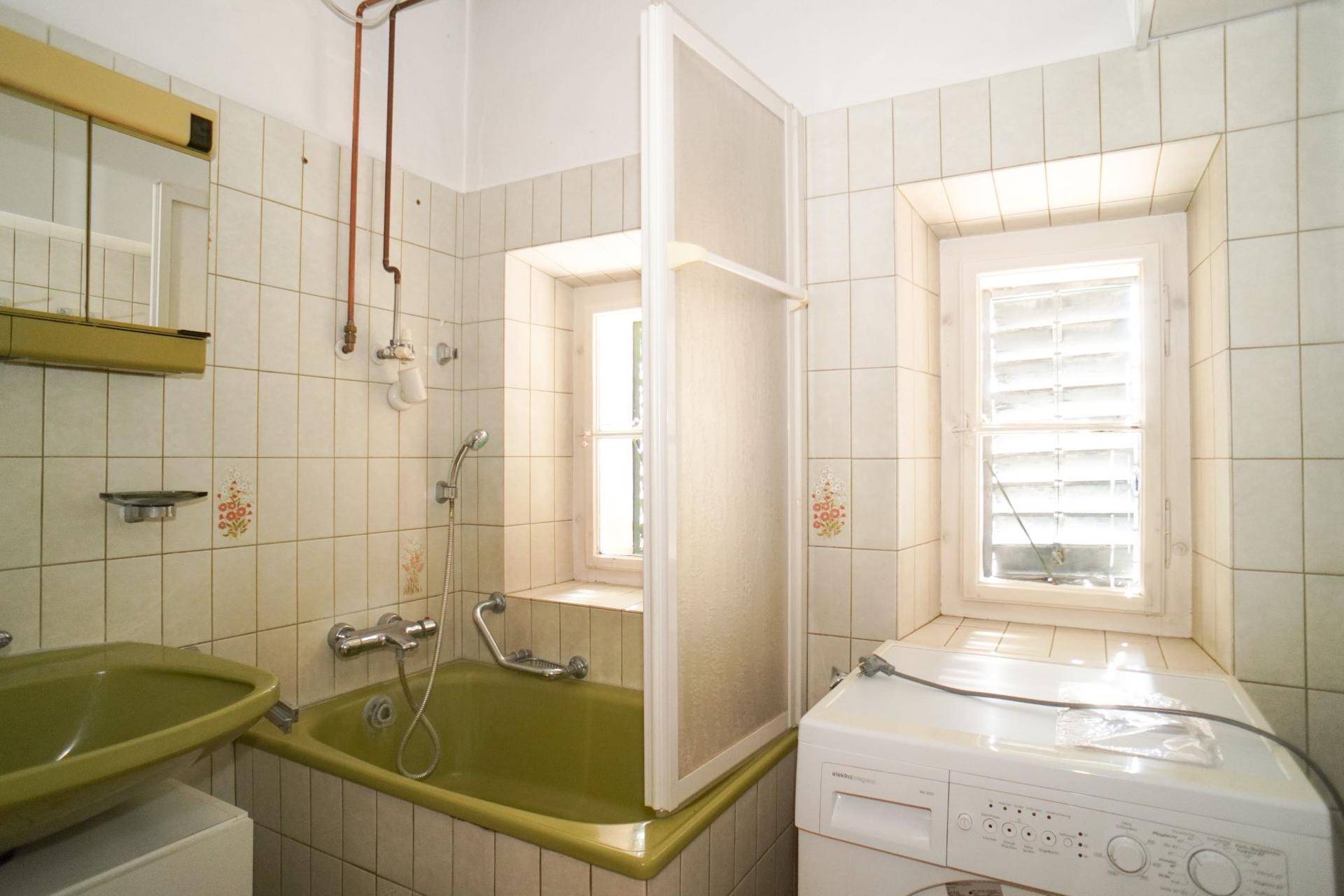 Badezimmer im 80er-Jahre-Stil mit grüner Badewanne, Fliesen und zwei Fenstern.