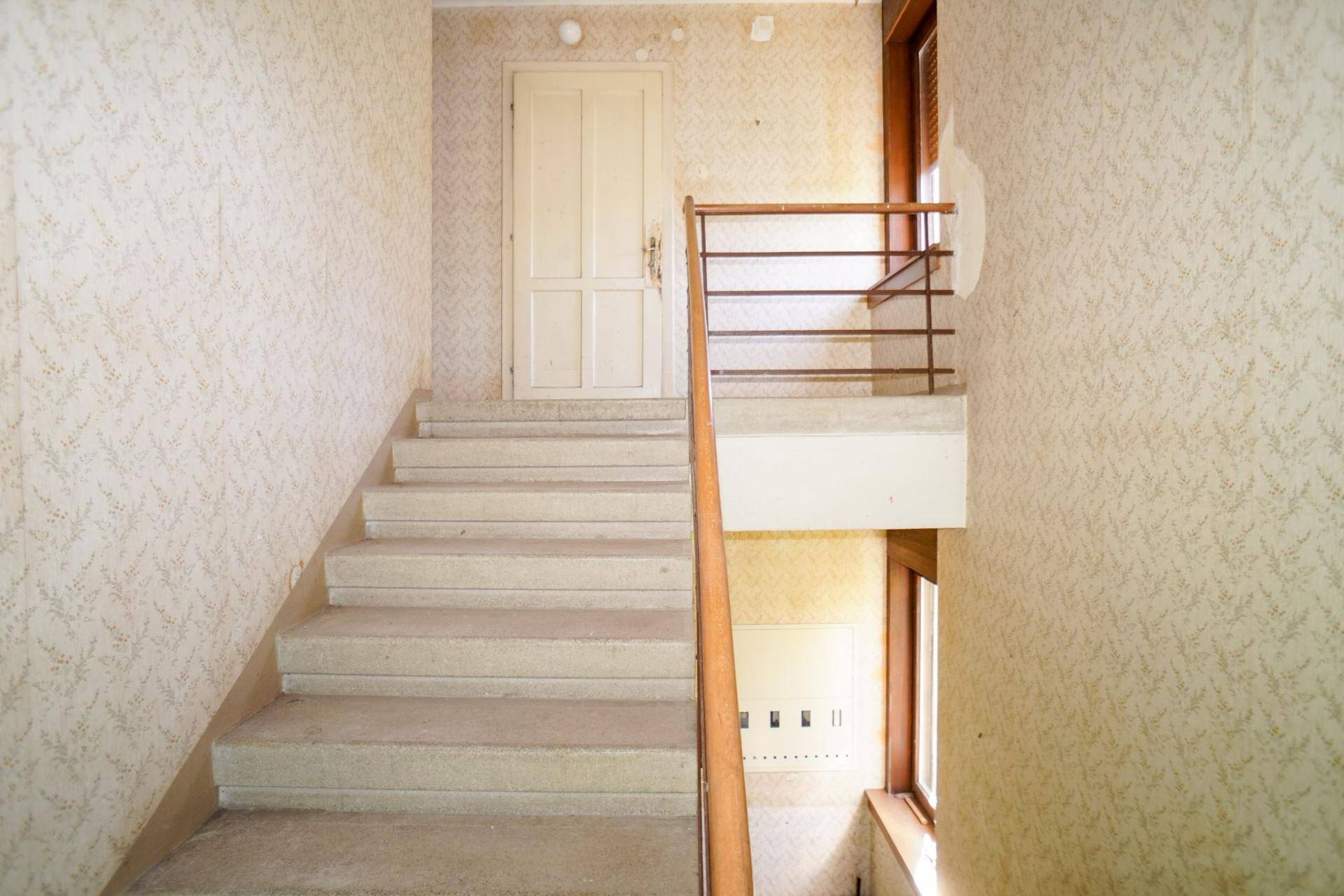 Treppenhaus mit Steinstufen, Holzgeländer und gemusterter Tapete, die Renovierung benötigt.