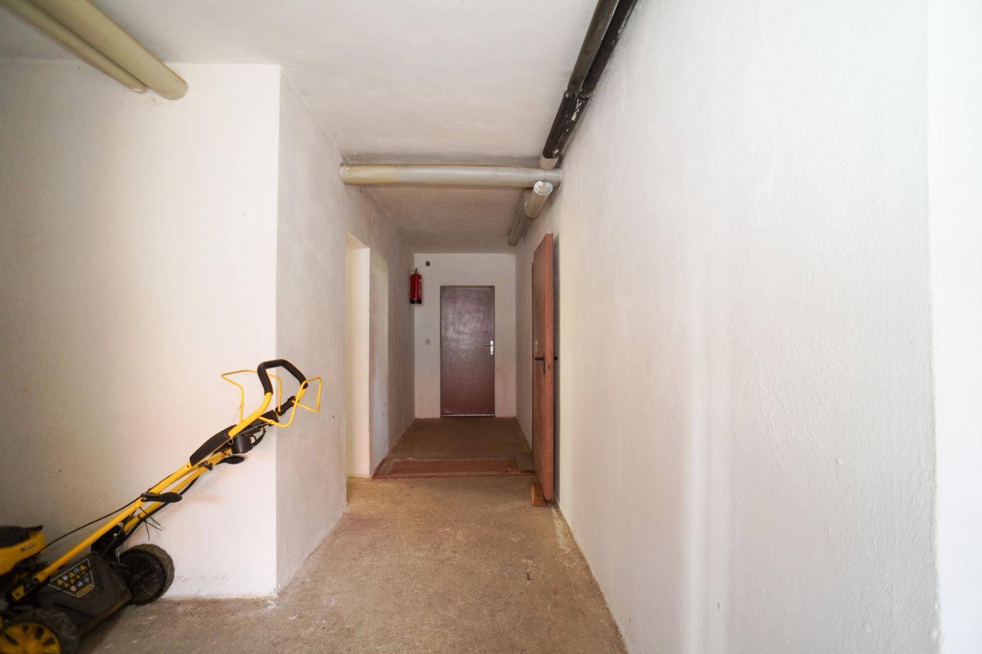Langer Kellerflur mit Betonboden, weiß gestrichenen Wänden und einer Rasenmäher.