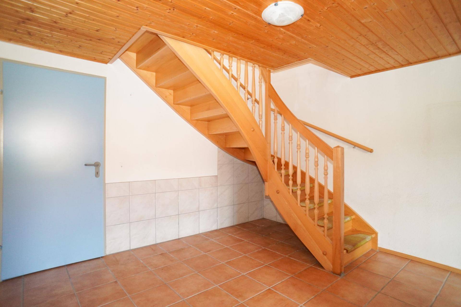 Heller Flur im Untergeschoss mit Holztreppe, Fliesenboden und einer blauen Tür.