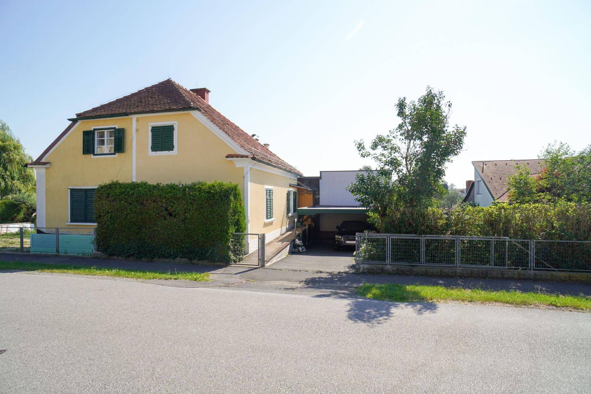 Gelbes Einfamilienhaus mit grünen Fensterläden und Carport, von der Straße aus gesehen.