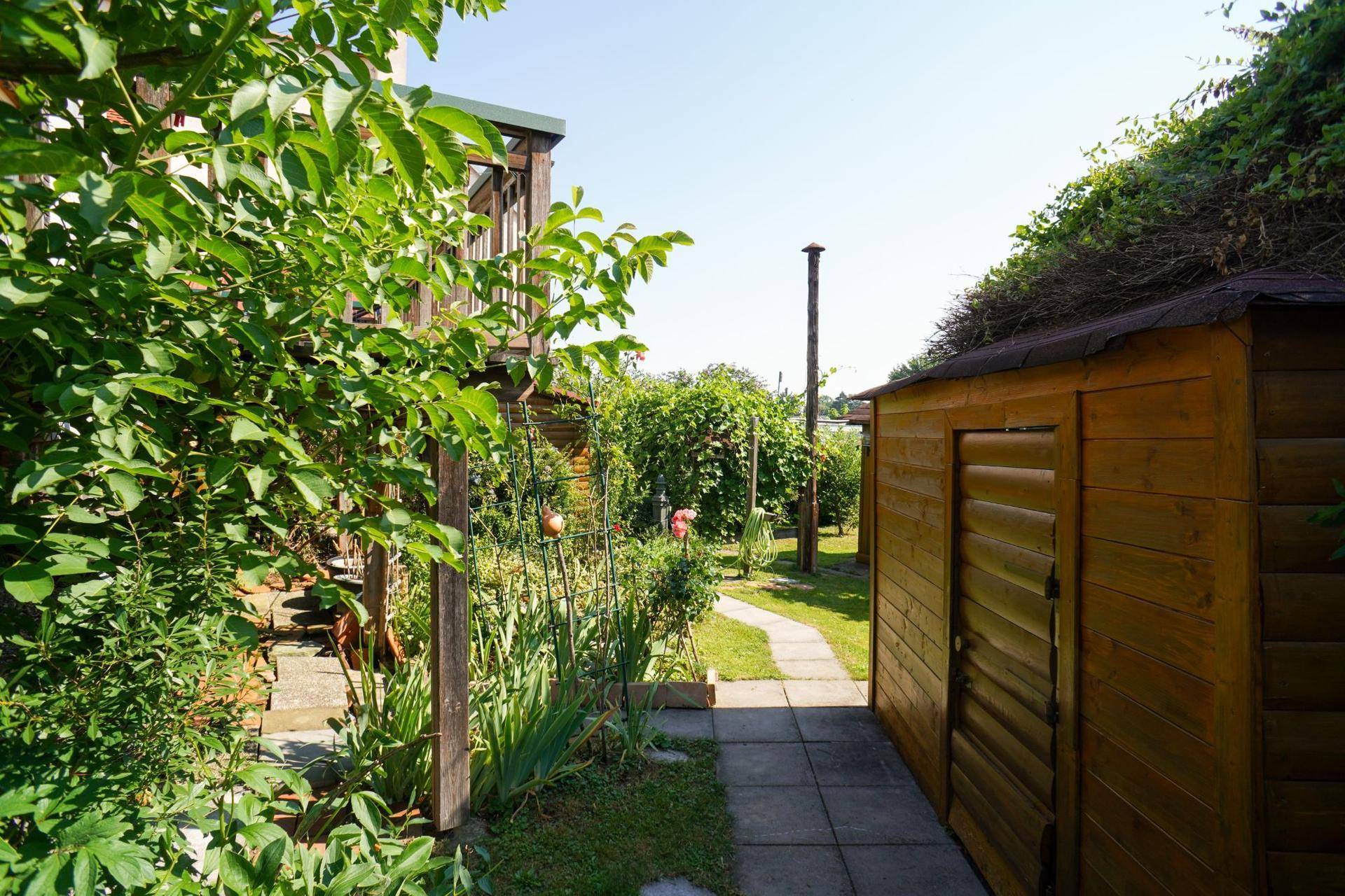 Seitlicher Blick auf den Garten mit Gartenhaus, üppiger Bepflanzung und dem Hauptgebäude im Hintergrund.