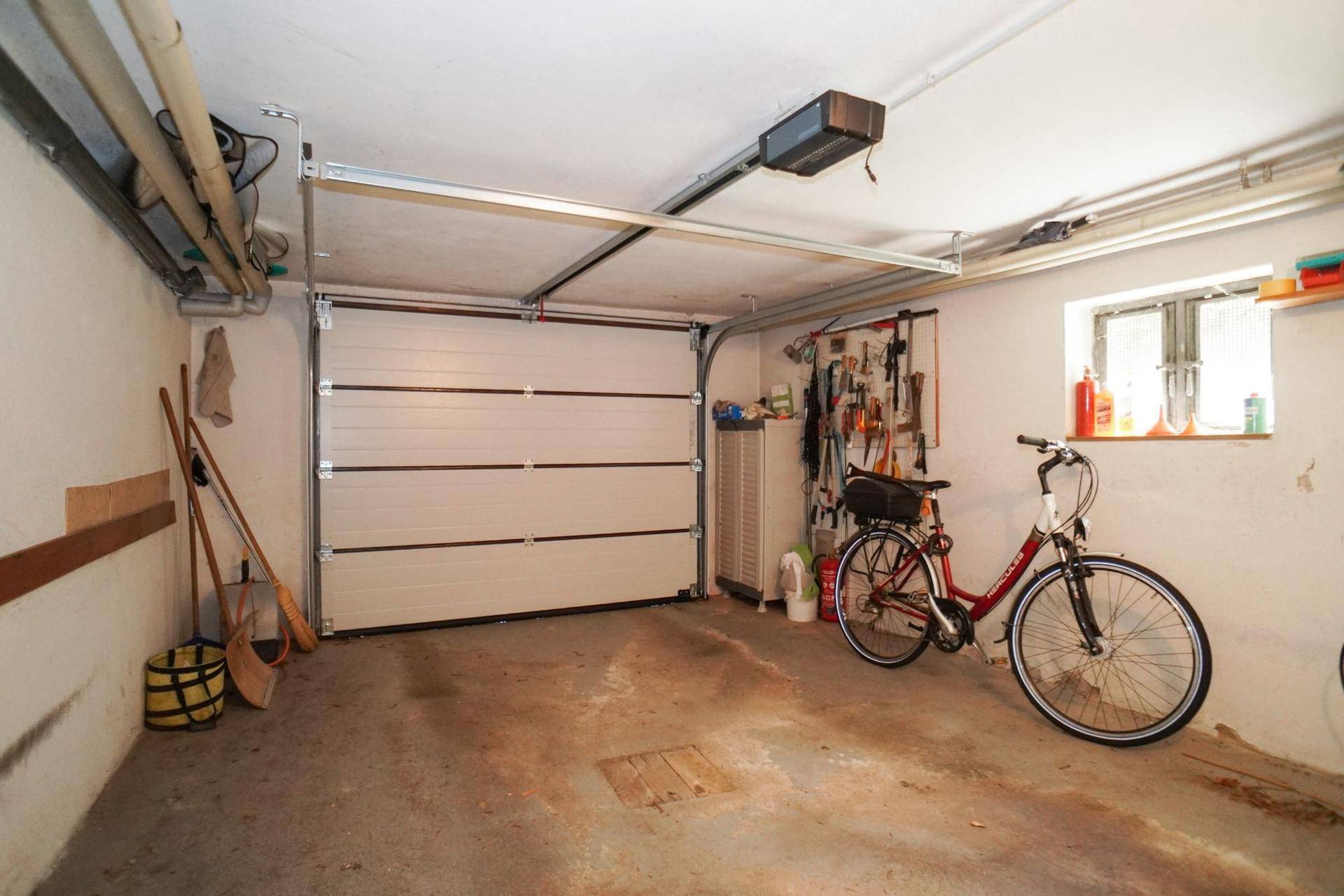 Geräumige Garage mit Sektionaltor, Werkzeugwand und Fenster, bietet Platz für Fahrzeug und Fahrräder.