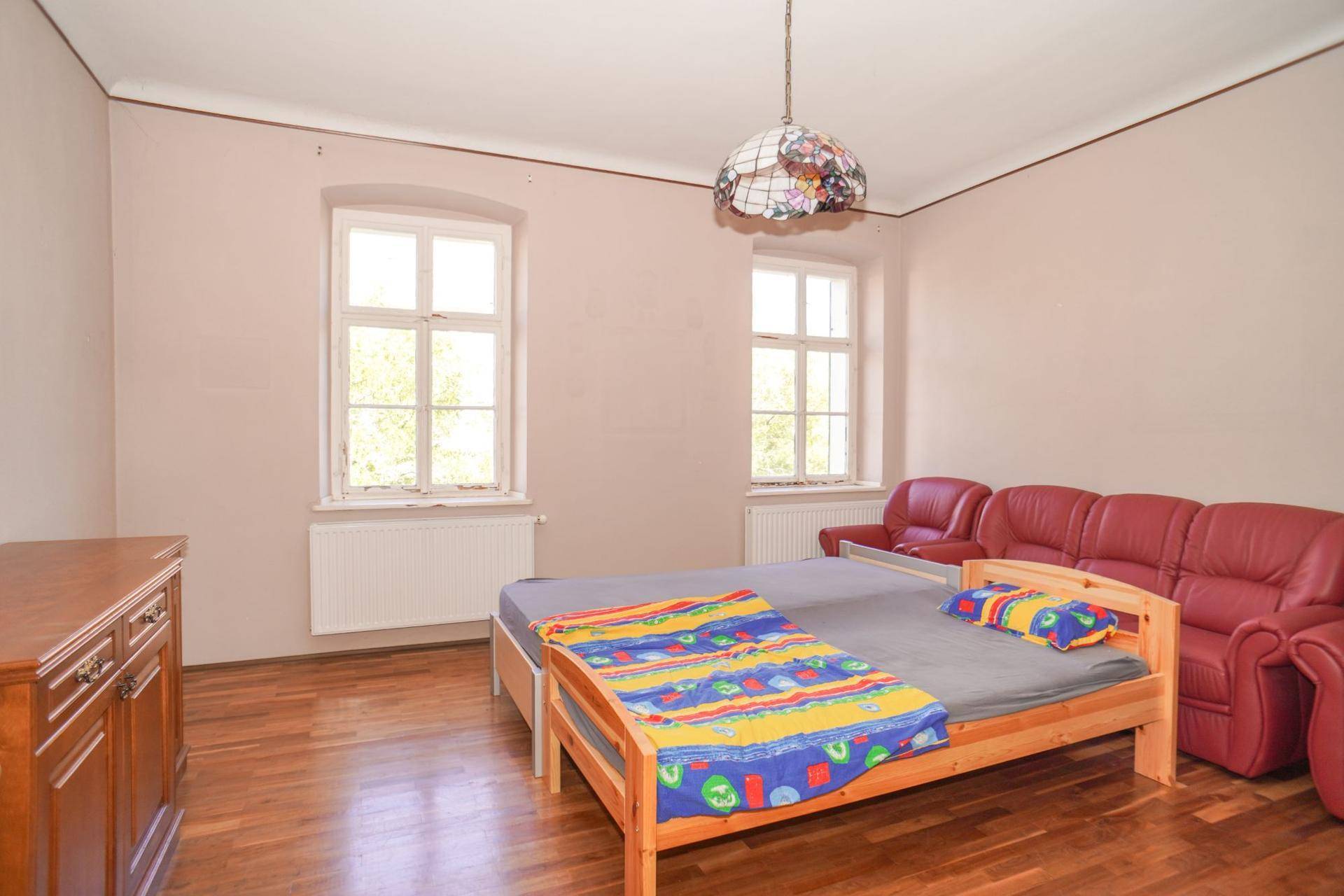 Helles Schlafzimmer mit Holzboden, einem Bett und rotem Sofa, durchflutet von Tageslicht.