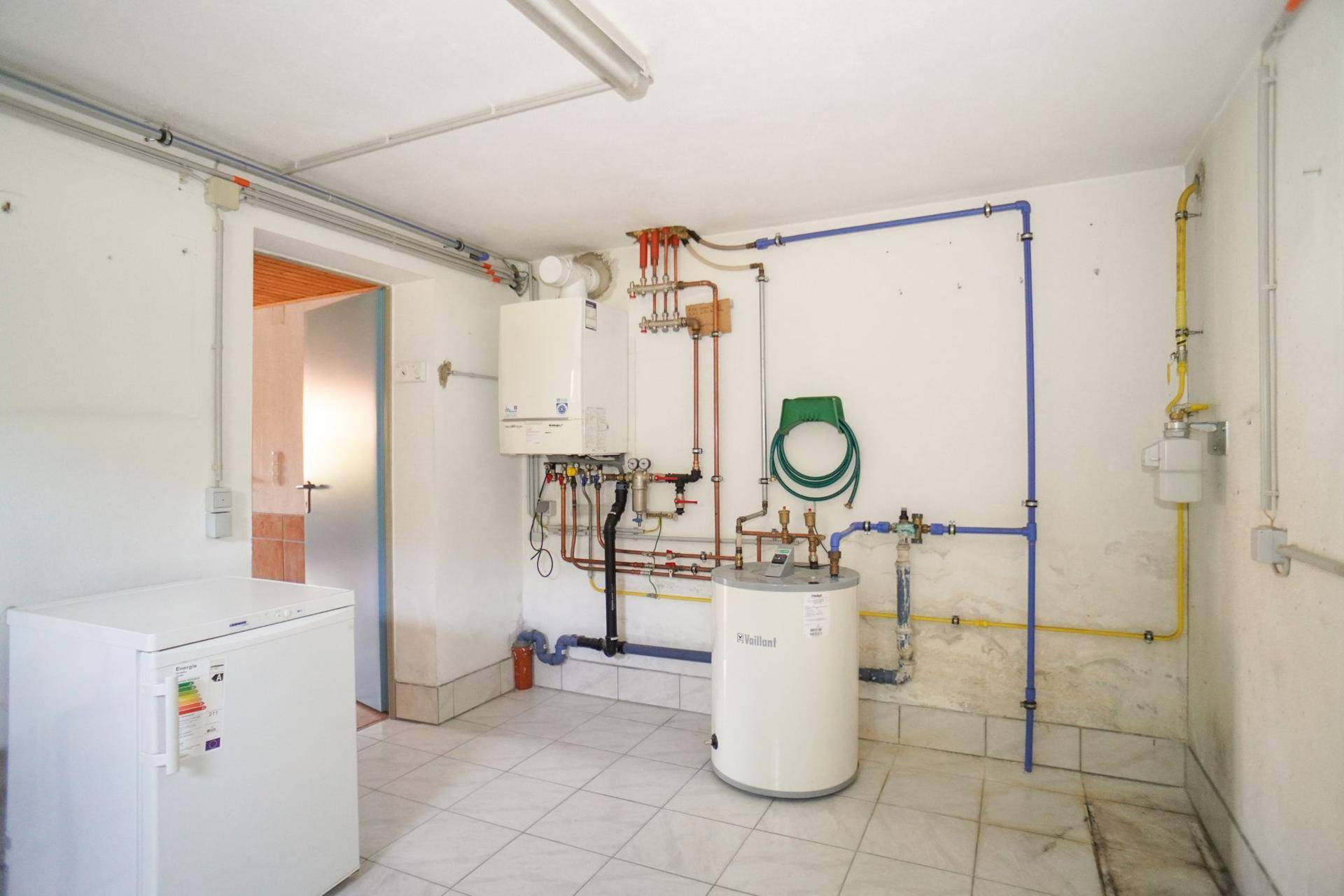 Heizungsraum mit Warmwasserboiler, Heizungsanlage und freiliegenden Leitungen sowie einem Gefrierschrank.