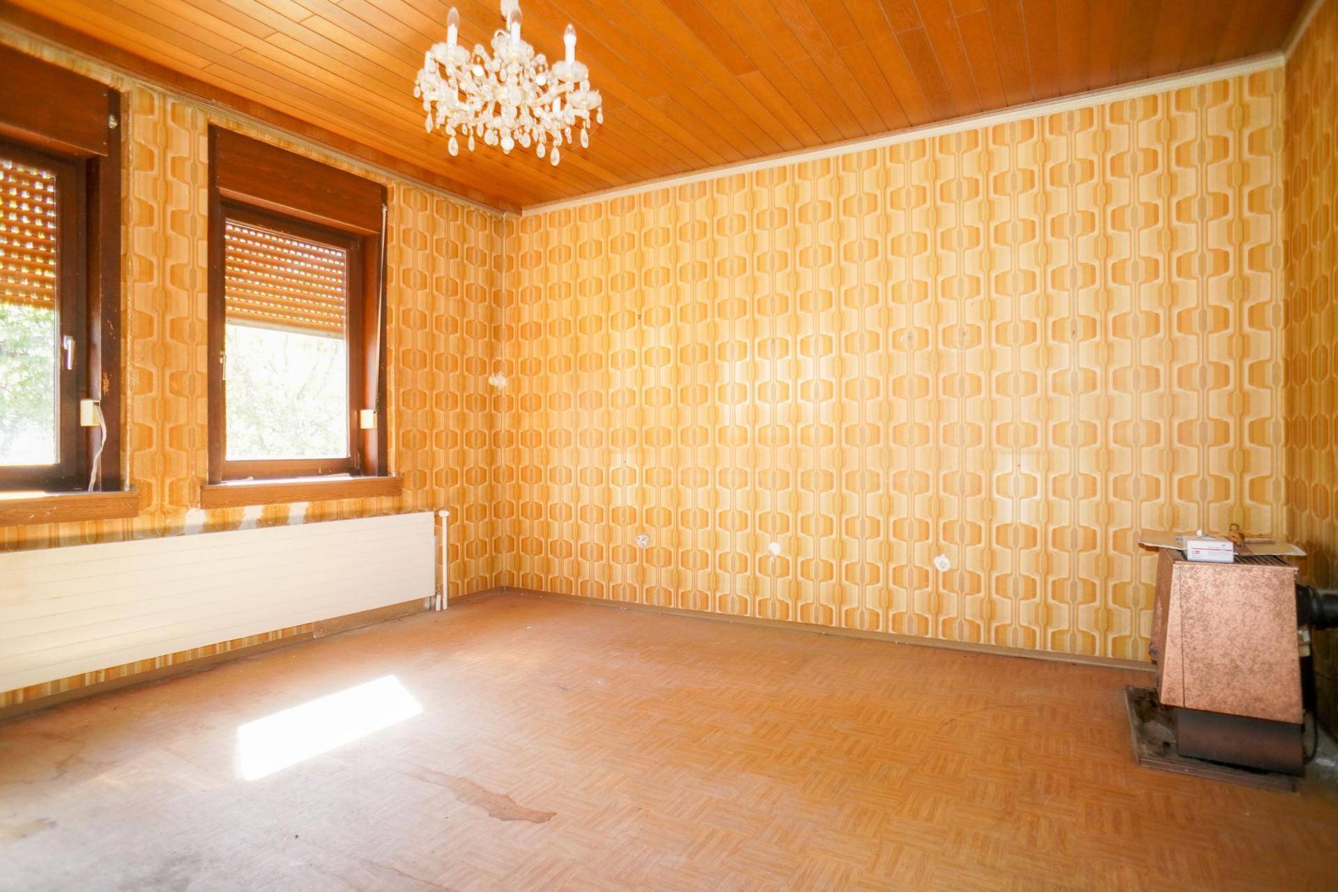 Wohnzimmer mit gemusterter Tapete, Holzdecke und einem alten Ofen, der Renovierung benötigt.
