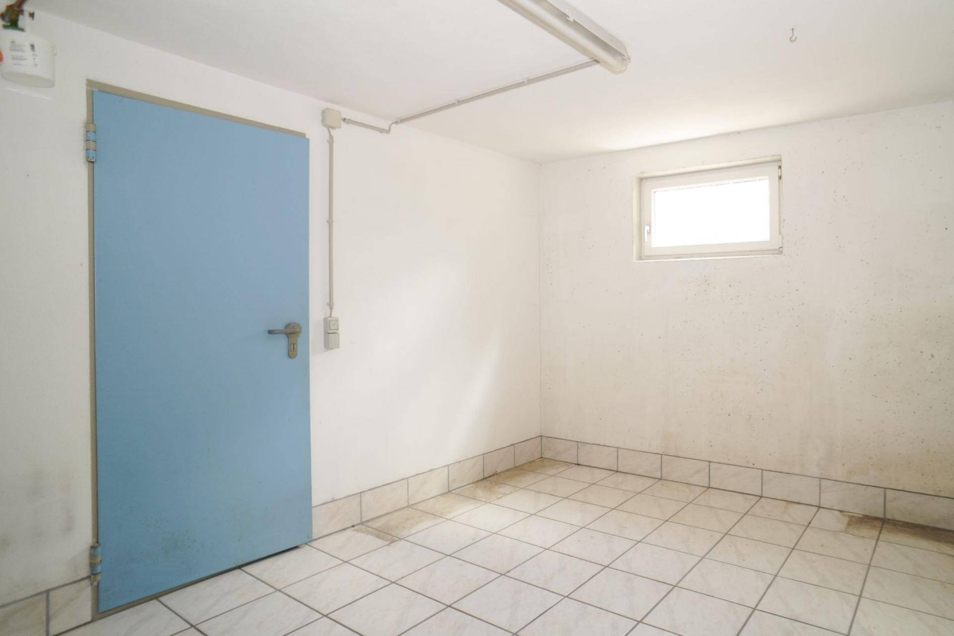 Leerer Kellerraum mit Fliesenboden, einer blauen Tür und einem kleinen Fenster für Tageslicht.