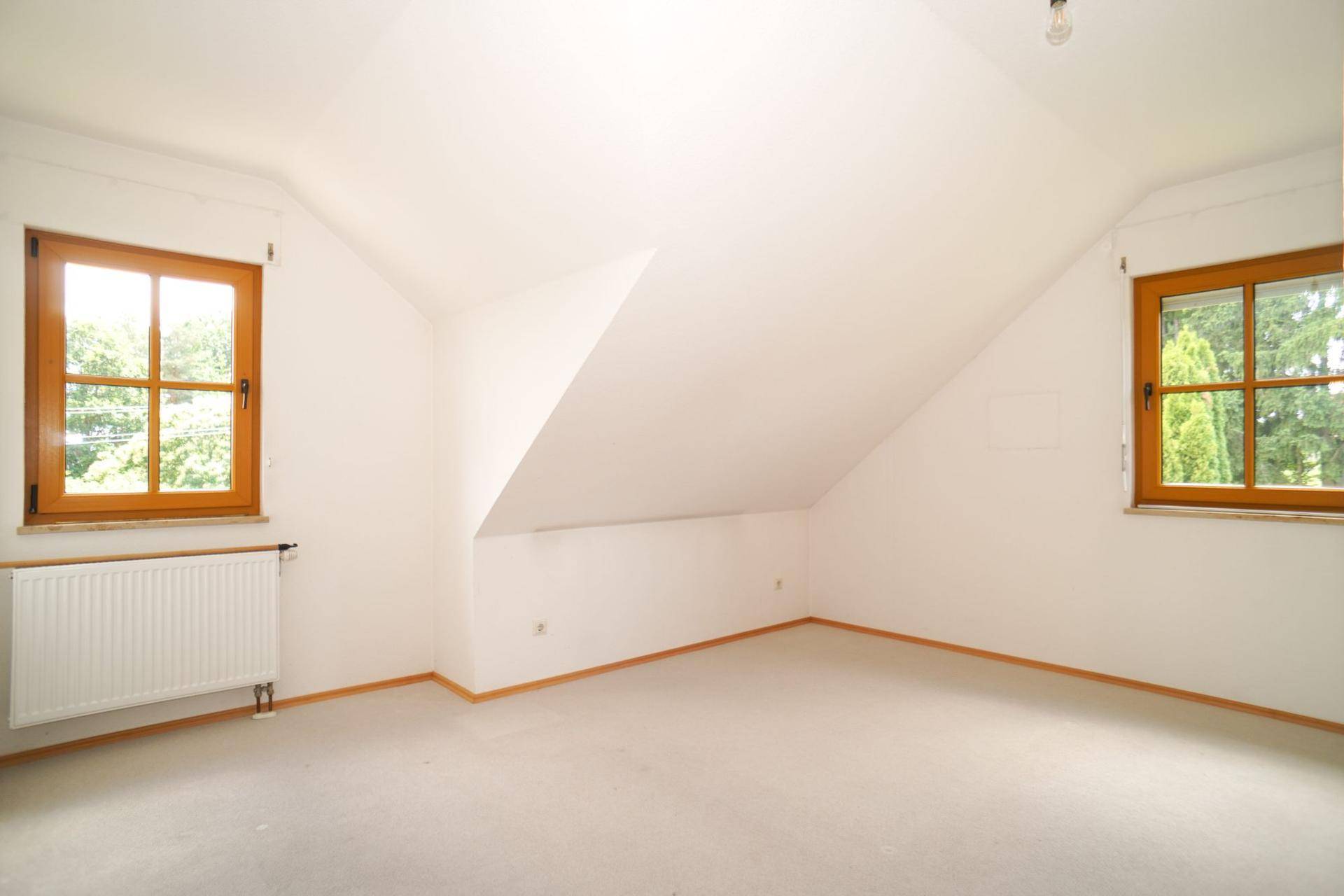 Geräumiges Dachgeschoss-Schlafzimmer mit Teppichboden, Dachschräge und zwei Fenstern mit grüner Aussicht.