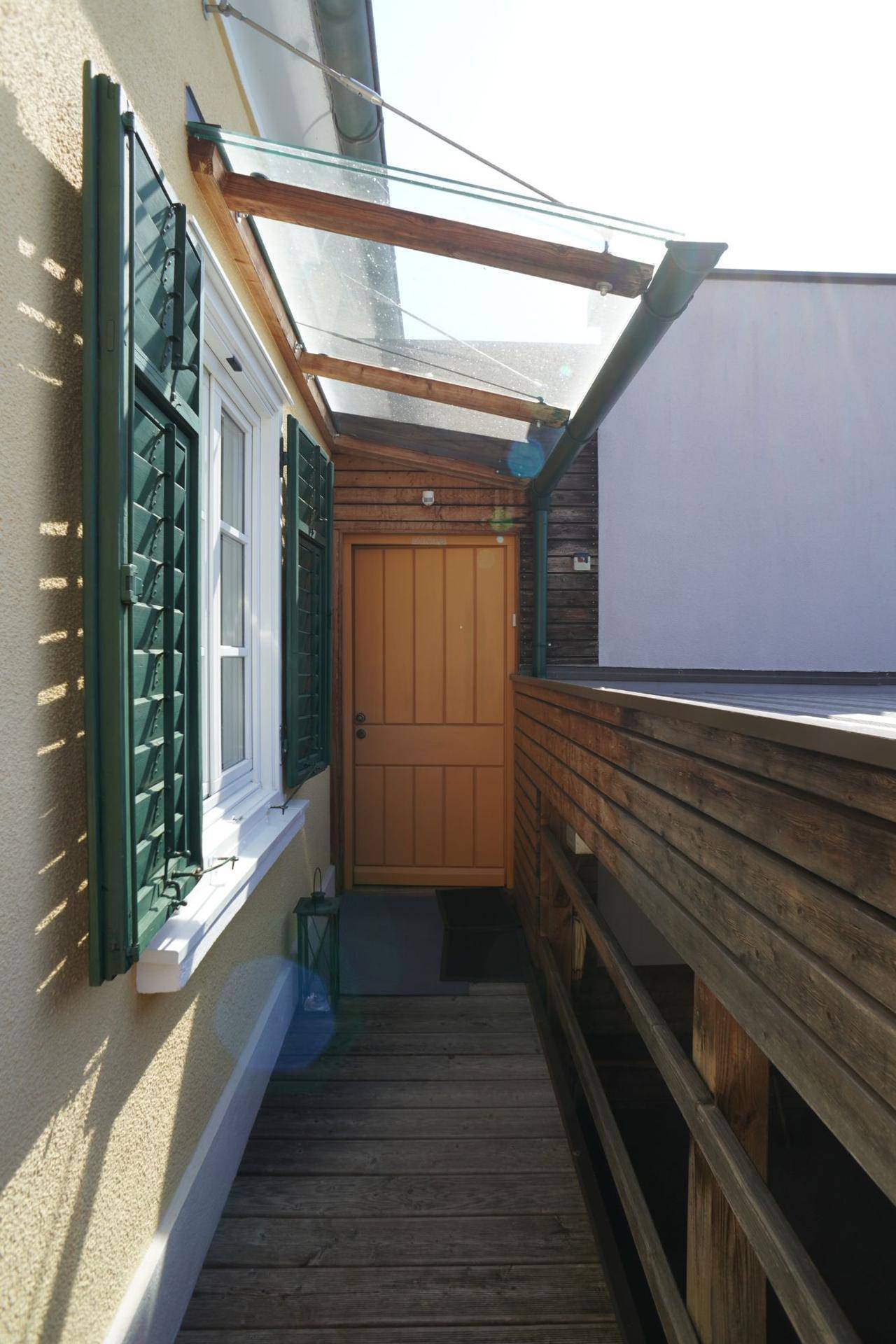 Überdachter Eingangsbereich mit Holzgeländer und Fensterläden an der sonnigen Hausfassade.