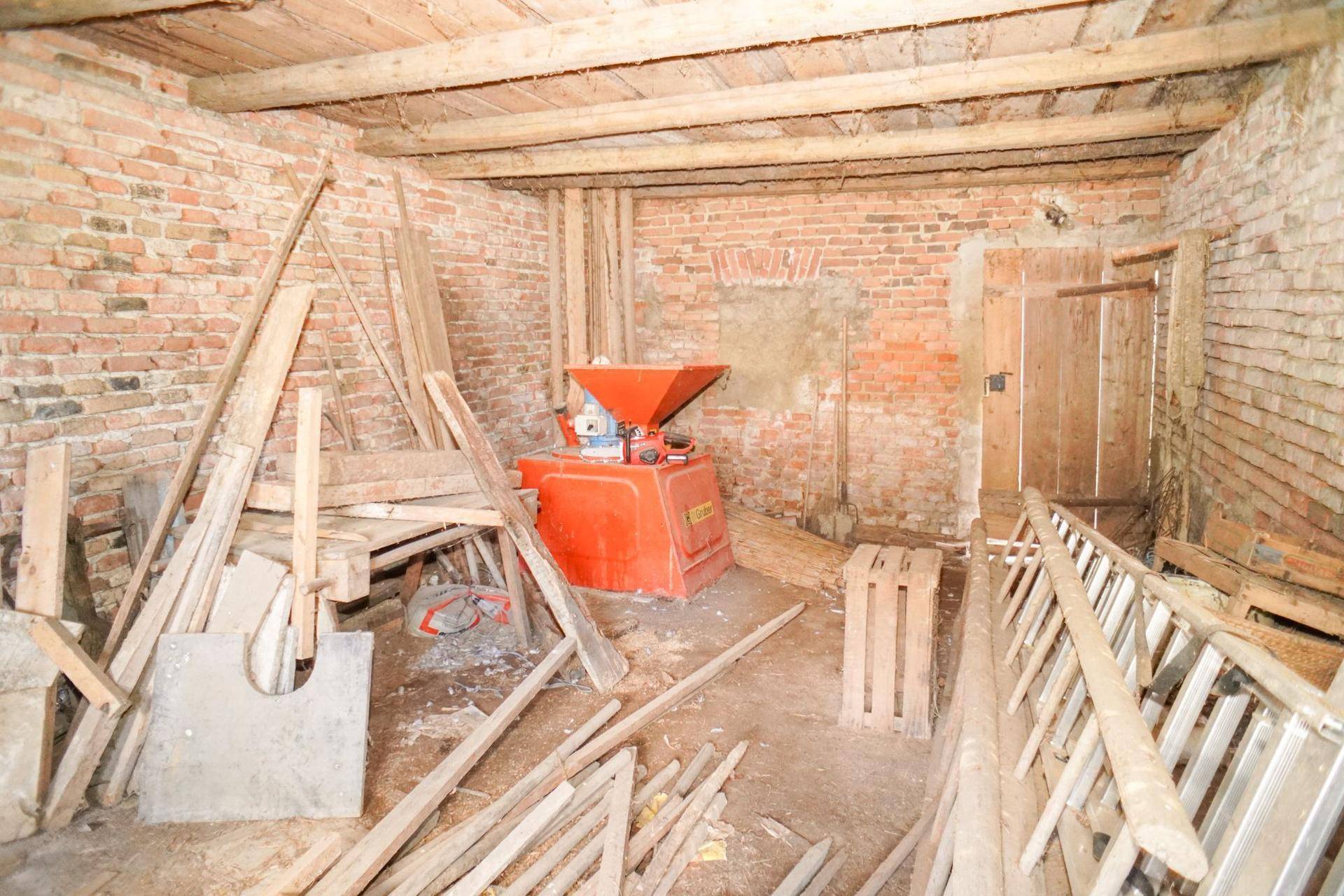 Werkstattraum mit unverputzten Ziegelwänden, Holzbalken und einer roten Maschine, voller Baumaterialien.