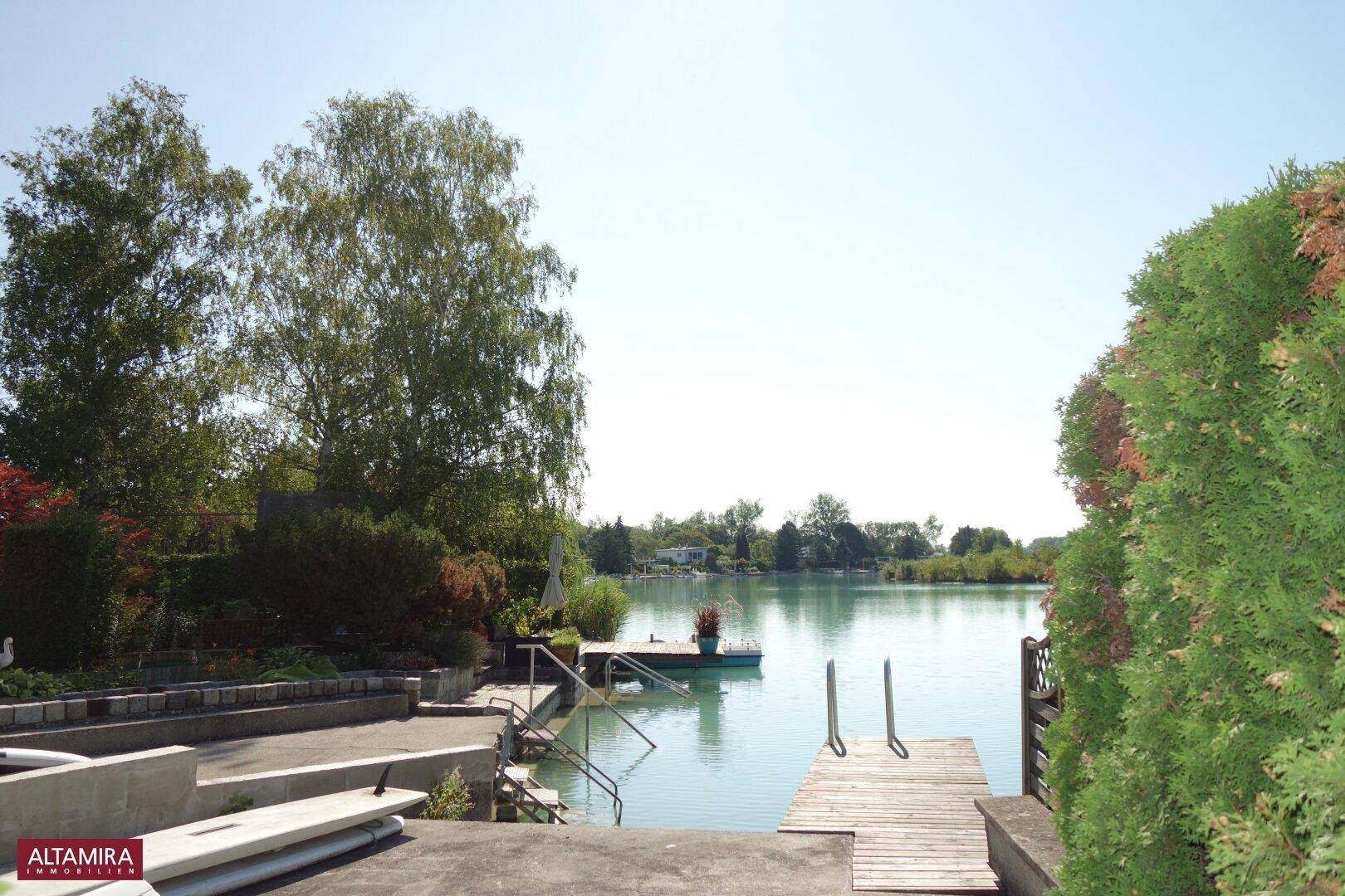 Panoramablick auf den See mit Holzsteg und Treppen, umgeben von grüner Vegetation.